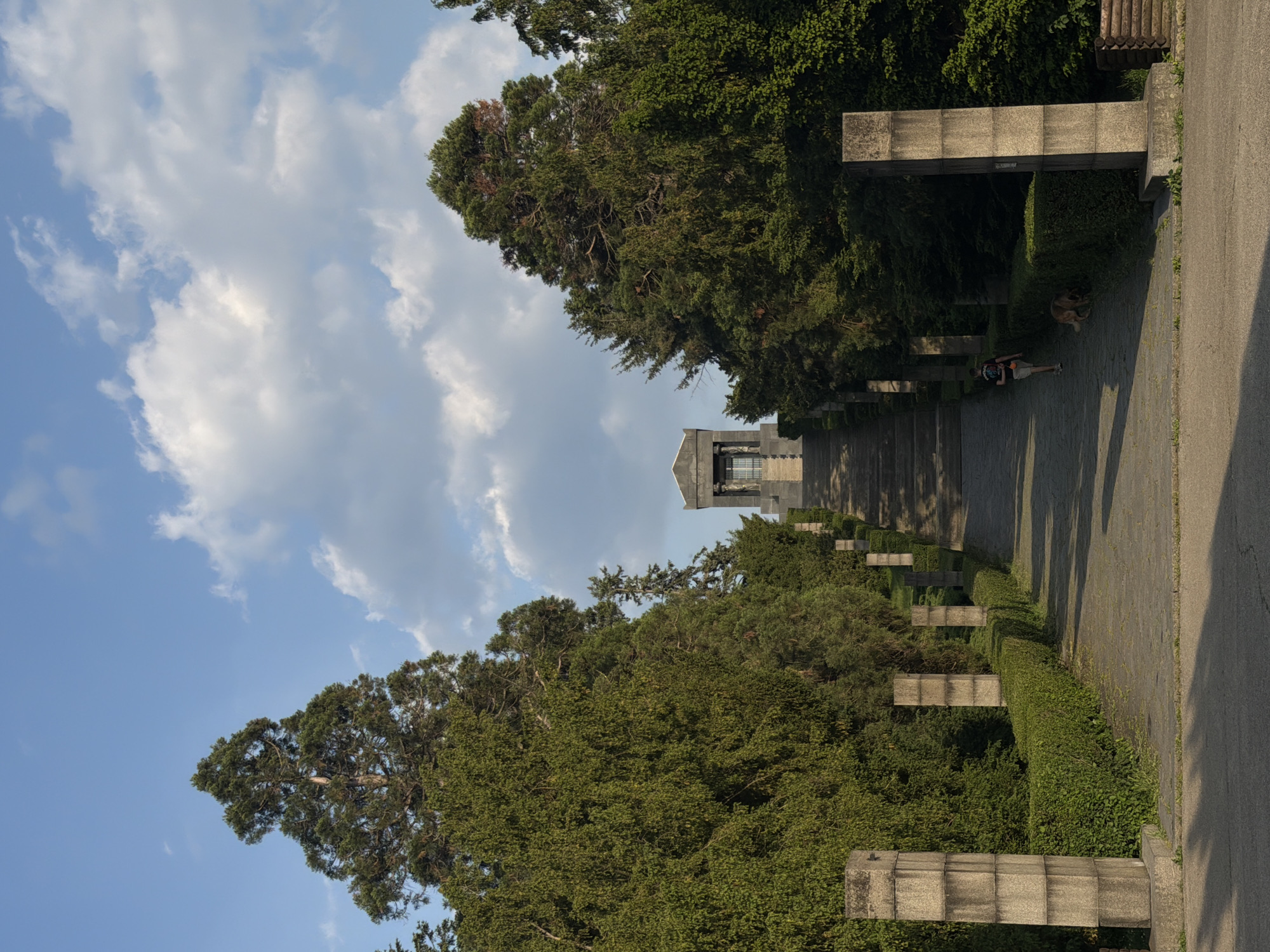 Captured in Vozdovac Urban Municipality, Serbia, the image presents a scene along a stone path lined with manicured hedges and concrete structures. The path leads towards a building with a tower in the distance. A person and a dog are resting on the path to the right. The sky, filled with clouds, frames the upper left portion of the image, contrasting with the dense greenery and architecture.