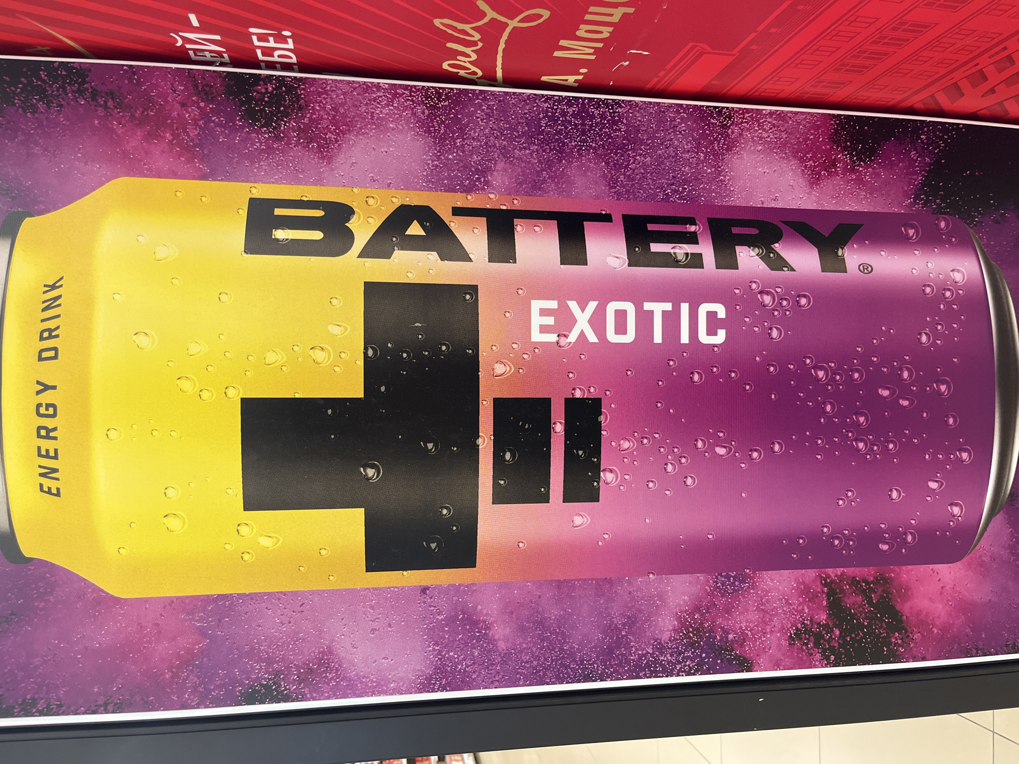 The image shows a close-up of a large advertisement for "BATTERY ENERGY DRINK" on what appears to be a wall or display panel. The advertisement features a stylized, yellow and pink can with water droplets, and black lettering. The words "ENERGY DRINK" are at the top of the can, and "BATTERY" is written vertically down the right side. "EXOTIC" is written vertically on the left side of the can. To the right of the advertisement, a portion of a red background with gold lettering is visible, with the text "БЕ!" and what appears to be a signature and name starting with "А. Мац". The advertisement is framed by a black border on the left. The overall impression is of a promotional display, likely in an indoor public space. The location in Kharkiv, Ukraine is not discernible from the image itself.