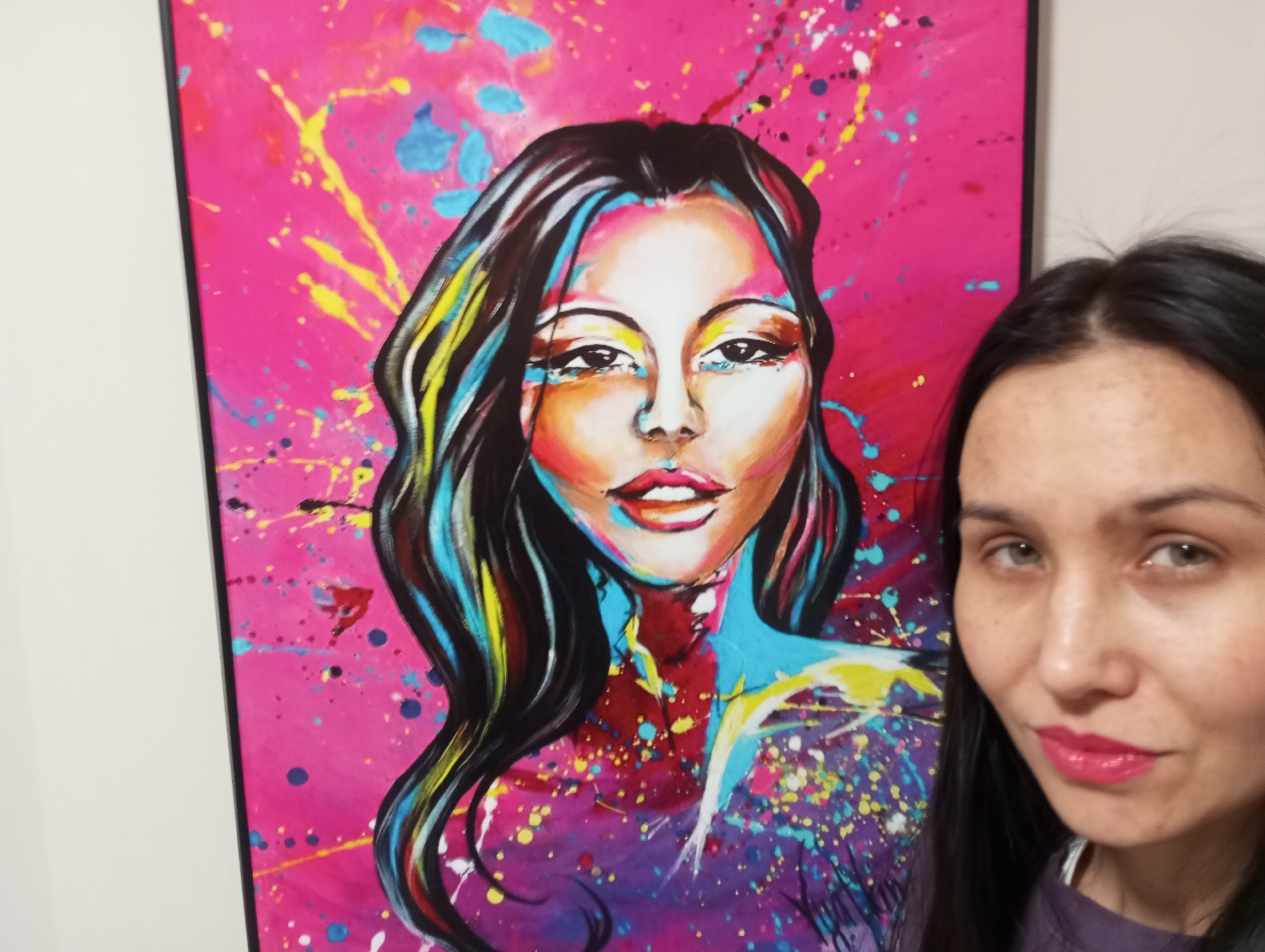 A woman is taking a selfie with a portrait. The portrait is a brightly colored painting of a woman with dark hair, a light complexion, and red lips. The background of the painting is a splash of pink, blue, yellow, and white paint. The woman taking the selfie has long, dark hair and is wearing a purple shirt. The background is a white wall. There is no indication of location, nor any basis to conclude the scene takes place in Pervomaysky, Russia.