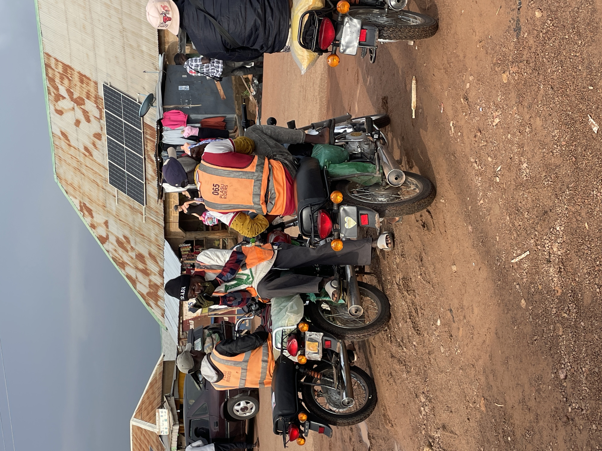 The scene depicts a dirt road in what appears to be a rural or semi-rural area. Several motorcycles are parked in a row, each appearing to be loaded with goods or supplies. The riders are wearing either casual clothing or vests. Buildings line one side of the road, one has a solar panel installed on its corrugated metal roof. The sky is overcast, suggesting a cloudy or potentially rainy day.