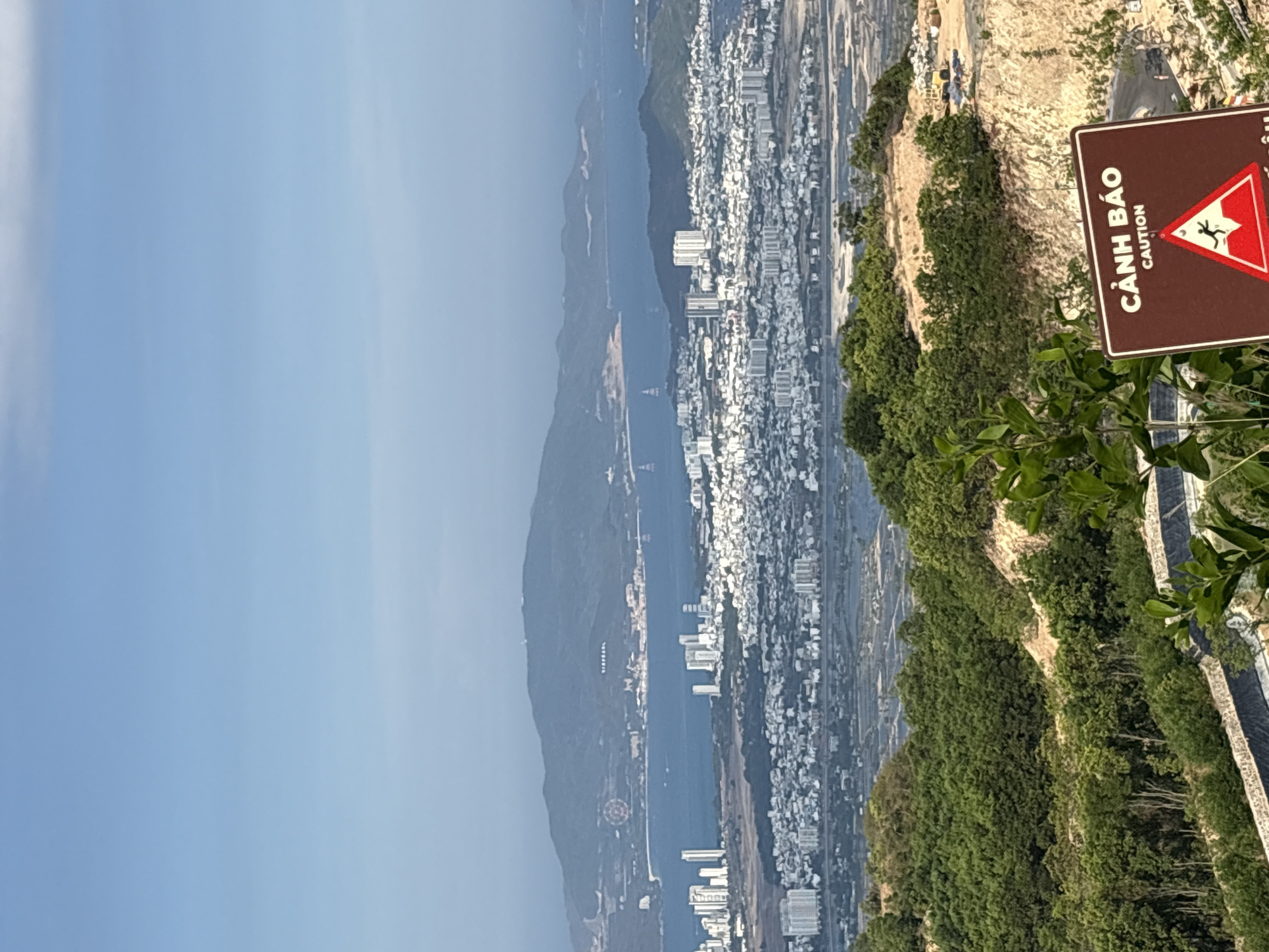 An aerial view shows Tay Nha Trang Ward, Vietnam, along the coast and bay, observed from a hillside perspective. The clear blue sky contrasts with the cityscape with numerous white buildings extending lengthwise along the coastal strip. A road parallels the buildings. The bay features a distant mountain range. Prominently positioned on the right foreground is a brown caution sign with a red warning triangle, marked with a stick figure falling, and Vietnamese text above the English word "Caution." A rugged cliff with shrubbery and a narrow paved pathway lines the foreground.