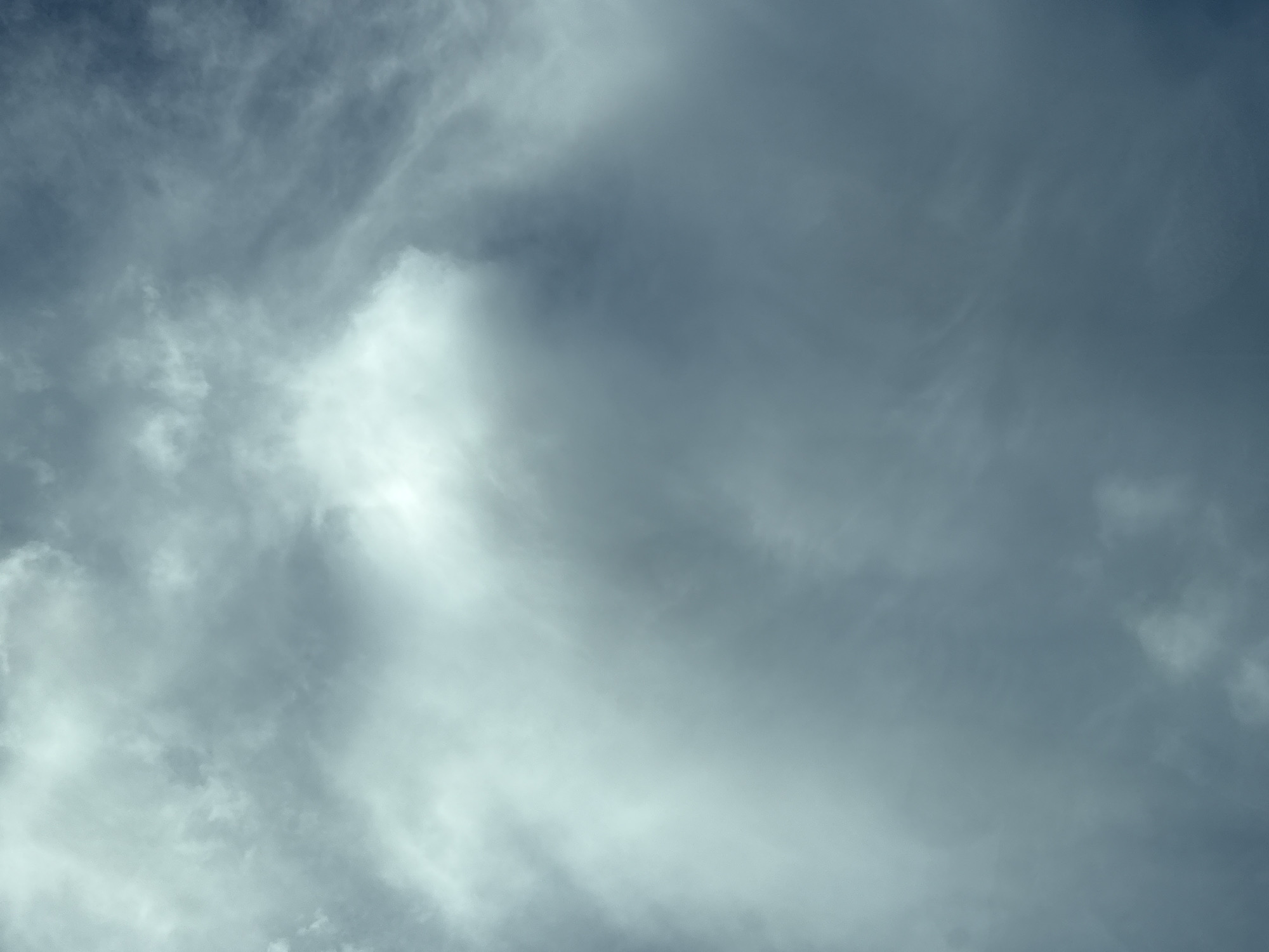 The image depicts a sky filled with dramatic, turbulent clouds. The lighting suggests either early morning or late afternoon, with the sun partially obscured, creating a sense of diffused, ethereal light. The colors are muted, dominated by shades of grey and white, with hints of blue in the darker areas of the sky. The texture of the clouds ranges from wispy and delicate to thick and voluminous, suggesting movement and atmospheric change. There are no discernible human subjects, objects, or specific landmarks visible that would allow for identification of a location within Wrocław, Poland. The scene is entirely focused on the natural phenomenon of the sky.