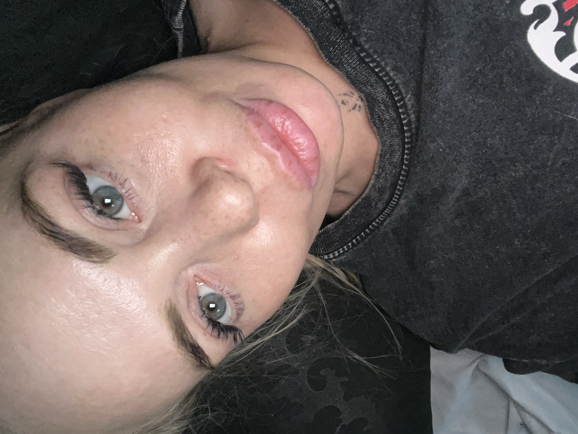 A close-up, high-angle, indoor selfie of a blonde woman's face is captured. She is wearing a dark grey t-shirt with a faded print. Her head is tilted to the left, and her eyes are looking slightly upwards and to the right. She has light blue eyes, long eyelashes, and lightly colored eyebrows. Her lips are full and pink. Her skin appears fair with some freckles. The background is dark and out of focus, suggesting a dimly lit indoor setting. There is a hint of light-colored fabric, possibly a pillow or blanket, in the bottom left of the frame. A faint tattoo is visible on her neck. The lighting in the image is soft and appears to be coming from above and slightly to the front.