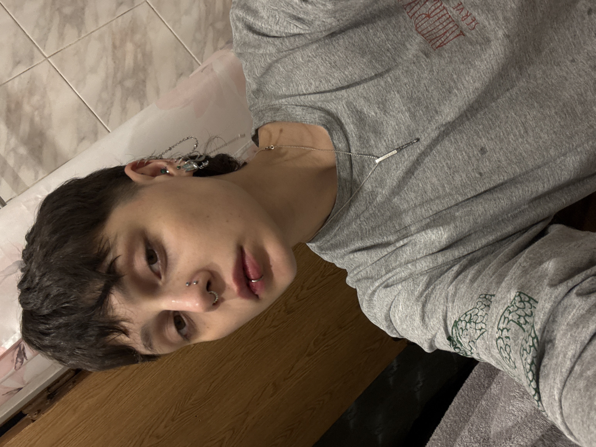 A young person with short, dark hair is lying down. They have a nose ring and lip ring. The person is wearing a grey long-sleeved shirt. The shirt has an inscription in red on the upper left chest and an image of foliage in green on the left sleeve. They are wearing jewelry in their ear and a necklace. The background includes a tiled wall, what appears to be a bathtub, and a wooden surface. There is no evidence to suggest the location of Saint Petersburg, Russia.
