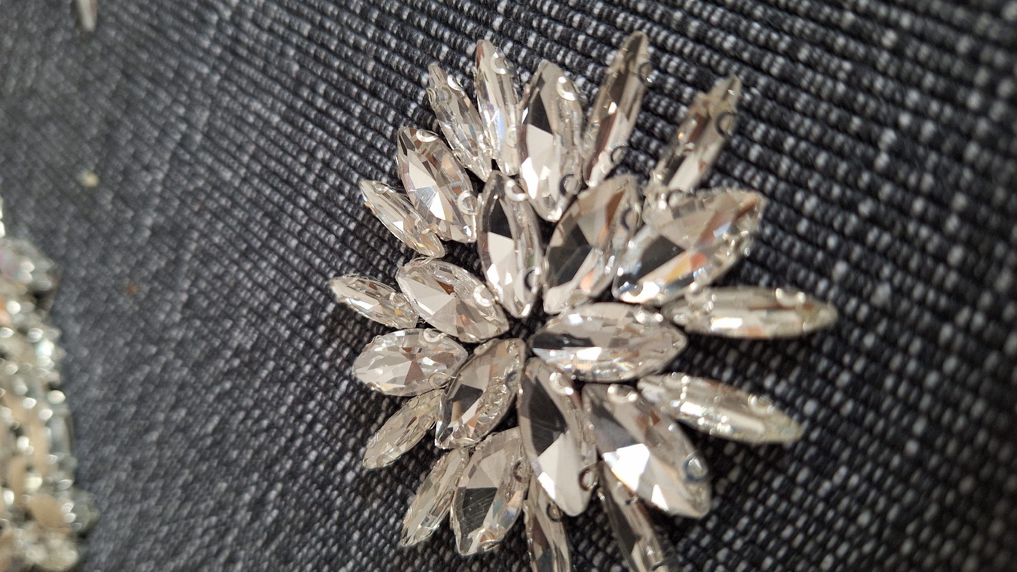The image shows a close-up of a denim fabric adorned with an array of clear, faceted crystals or rhinestones. The crystals are arranged in a radial pattern, resembling a flower or starburst design. The denim fabric provides a textured, dark gray background that contrasts with the sparkling crystals. The crystals are tear-drop shaped with a slightly raised, rounded surface to catch the light. The location in Blagoevgrad, Bulgaria cannot be confirmed based on the image alone.