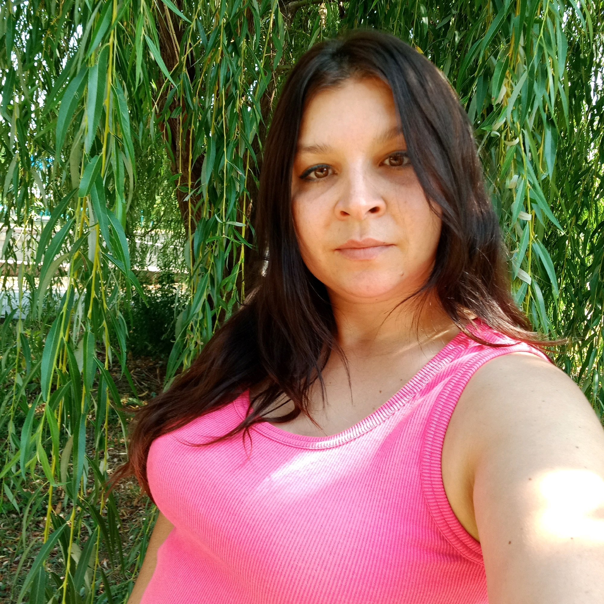 A woman with dark hair and brown eyes is pictured outdoors, with willow tree branches hanging in the background. She is wearing a pink ribbed tank top. The lighting suggests it is daytime. The scene appears to be a park or garden area, consistent with locations found within Kropyvnytskyi, Ukraine.