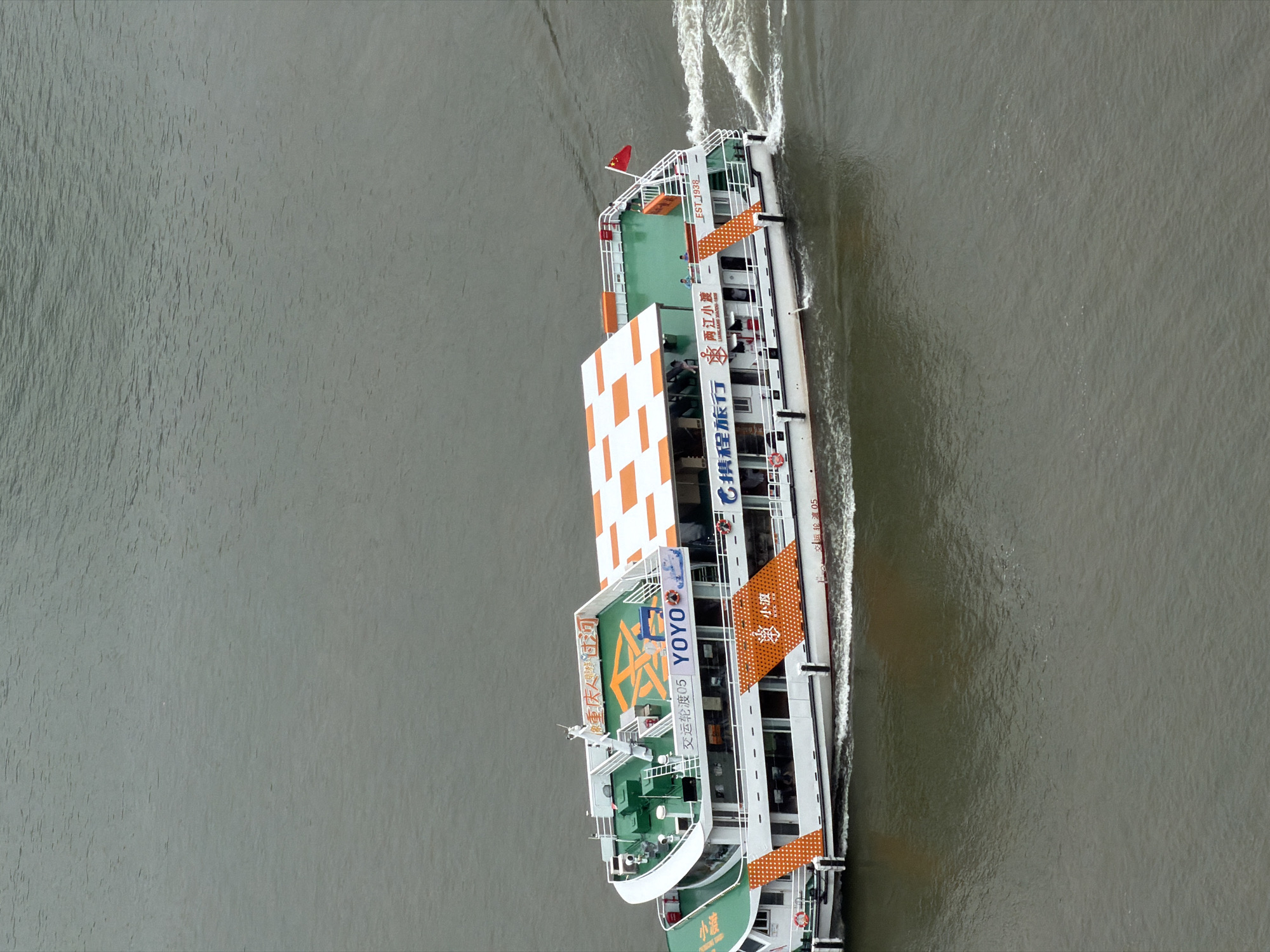 A white and green passenger ferry, identified by the Chinese characters and "YOYO" printed on its side, navigates through murky, choppy water. The ferry features a checkered orange and white roof and an orange pattern on its hull. A red Chinese flag flies from the stern. Several people are visible on the upper decks. The water suggests a river or bay, and the overall scene is overcast. The ferry is likely in Yuzhong District, China, given the context provided.