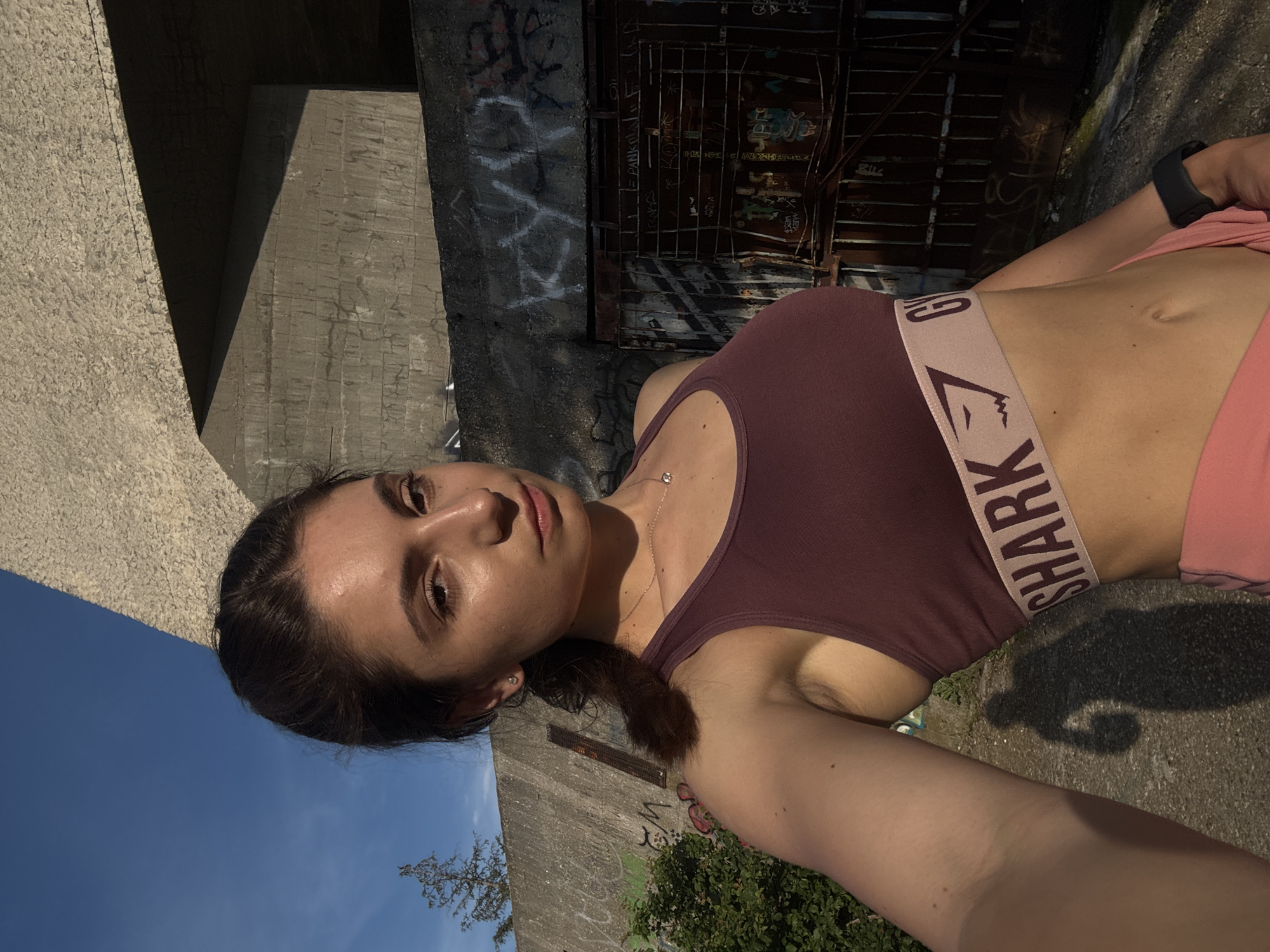 In Varna, Bulgaria, a young woman with dark hair pulled back in a ponytail takes a selfie outdoors. She is wearing a maroon sports bra with a pink band that reads "GYM SHARK" and pink shorts. The background consists of a concrete wall with graffiti, a rusted metal gate, and a clear blue sky. The sun is casting shadows on the ground and the woman's arm.