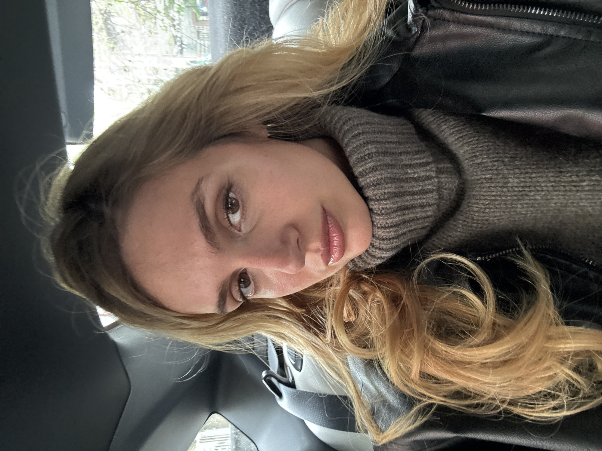 A young woman is depicted in a close-up, head-tilted selfie, positioned within the interior of a vehicle. She has long, wavy blonde hair with lighter highlights that frames her face and flows over her shoulders. Her features include fair skin, brown eyes, and full, glossy lips, with a neutral expression directed at the camera.
She is wearing a dark, ribbed turtleneck sweater, partially obscured by a black leather jacket featuring a visible zipper detail on the shoulder. A small hoop earring is visible on her right ear.
The background is blurred, showing a window view from the vehicle, revealing hints of bare tree branches against a bright, possibly overcast, daytime sky. The dark interior of the car, including parts of a seat or seatbelt, is discernible around her. Natural light from the window illuminates her face and hair. No text is visible in the image.
