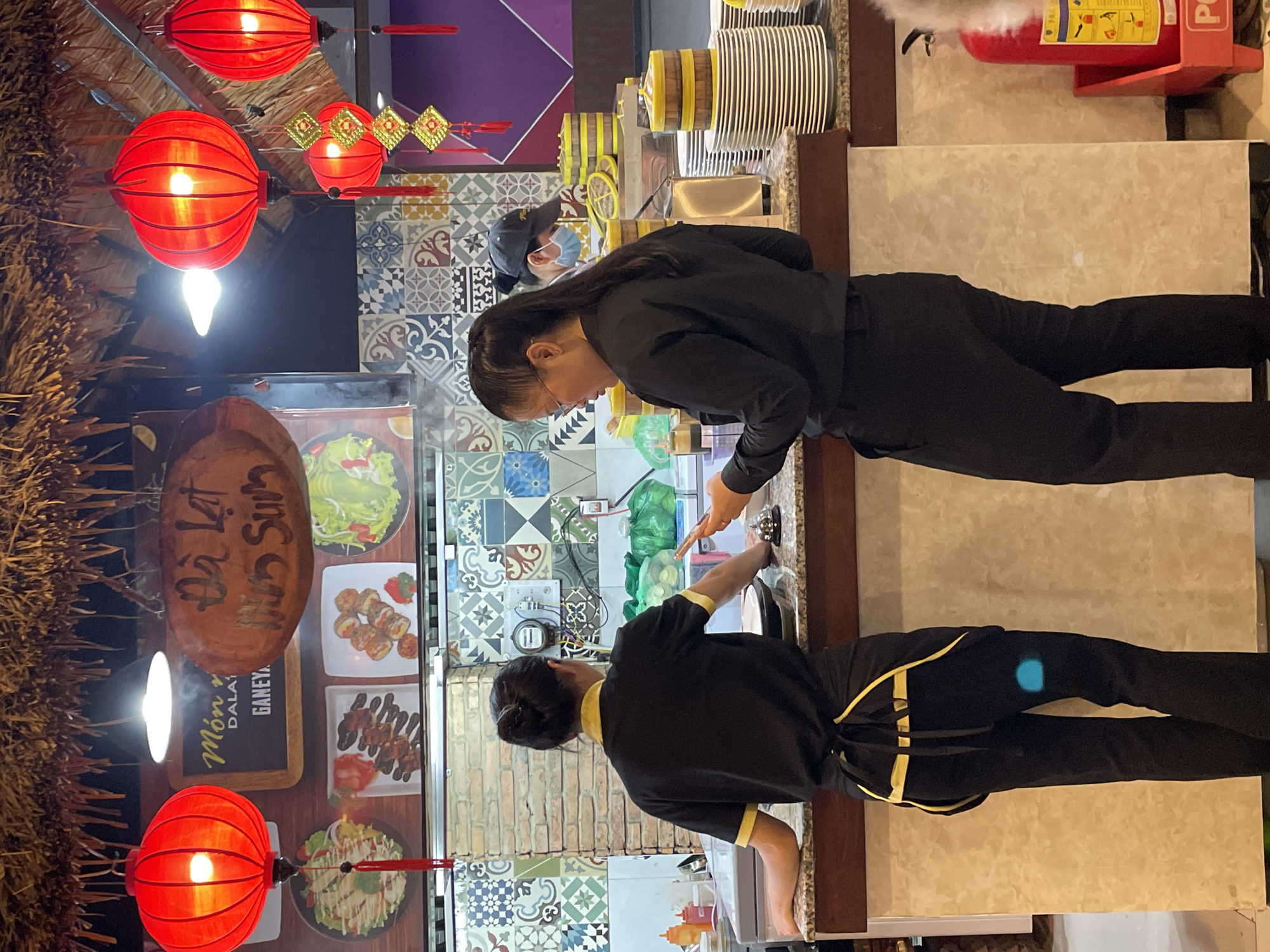 This image captures an indoor scene within a food establishment located in Da Lat, Vietnam, indicated by signs reading "Món ngon Đà Lạt" (Delicious Dishes Da Lat) and "Đà Lạt" on wooden plaques hanging from a thatched roof. The foreground features a granite-topped service counter behind which two female staff members are visible.
The staff member on the left is positioned with her back to the viewer, wearing a black short-sleeved uniform shirt with yellow trim on the collar and sleeves, and a black apron with yellow ties. Her dark hair is pulled back into a bun. Her left forearm rests on the counter, and her right hand appears to be near a black pan or bowl.
The staff member on the right is seen in profile, facing towards the right side of the frame. She wears a long-sleeved black shirt and black trousers, with her dark hair pulled back in a ponytail. She wears glasses and is actively looking down at and interacting with a white smartphone held in her hands. A small silver service bell is present on the counter between the two women.
Behind the counter, a patterned tiled wall with various geometric and floral motifs in shades of blue, green, brown, and white forms the background. A section of exposed brickwork is visible in the center of the wall. Several round food display boards show images of prepared dishes, including salads, skewers, and fried items. Above these displays, red spherical lanterns with internal lights and red tassels hang from the thatched ceiling, along with smaller gold and red decorative elements.
Further in the background, to the right, another person is partially visible behind a counter, wearing a dark cap and a light blue face mask, suggesting additional kitchen or service staff. Stacks of white plates and yellow-banded bamboo steam baskets are arranged on the counter. To the far right, a red fire extinguisher in a red stand is visible on the floor. Steam is faintly visible rising from behind the main counter on the left side of the frame.