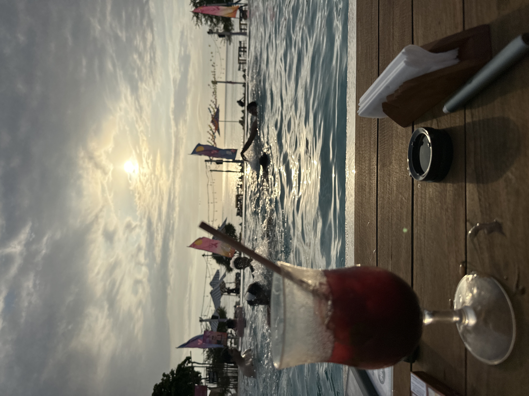 Here is a detailed description of the image:
**Location:** A swimming pool area in an unknown location in Indonesia. The presence of flags and the overall aesthetic suggest a resort or beach club setting.
**Environment:** The scene is captured during the daytime, with the sun visible through the clouds. The sky is partly cloudy, and the lighting is bright with some glare. The pool is filled with water, which reflects the sunlight and sky. The pool is surrounded by wooden decking.
**Objects:**
* A cocktail glass filled with a red drink, with condensation visible on the glass. A straw is also visible.
* A lens cap, a stylus, and a container with white napkins sit on the wooden decking next to the pool.
* Flags or banners are arranged along the poolside area, likely advertising the establishment or adding to the decorative atmosphere.
**People:**
* Several individuals are present in the pool, some closer to the edge and some further away.
* The people are swimming or standing in the water.
**Actions:**
* People are swimming or standing in the water.
* The photographer is capturing the scene, likely from a poolside location.
**Interactions:**
* The individuals in the pool are interacting with the water.
**Overall Impression:** The image presents a relaxing poolside scene, likely at a resort in Indonesia. The focus is on the cocktail and the pool, with people enjoying themselves in the water. The time of day and the lighting create a warm, inviting atmosphere.