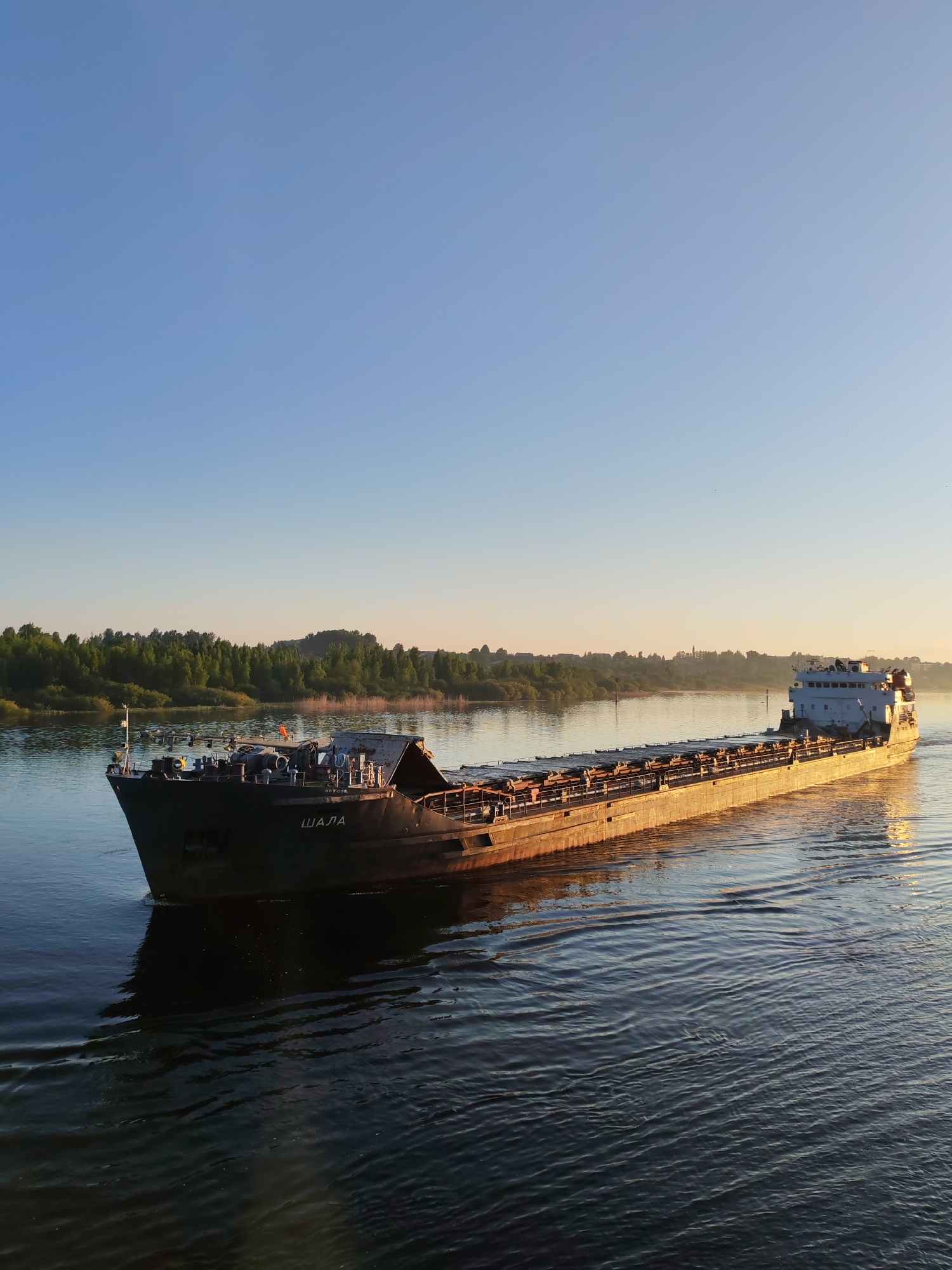 A cargo ship named "Shala" is traversing a body of water. The ship is loaded with cargo. The water is calm with small ripples. The sky is clear blue. The background shows a line of trees and vegetation along the water's edge. The image appears to be taken at sunrise or sunset, given the warm light. The location is Unknown, Unknown.