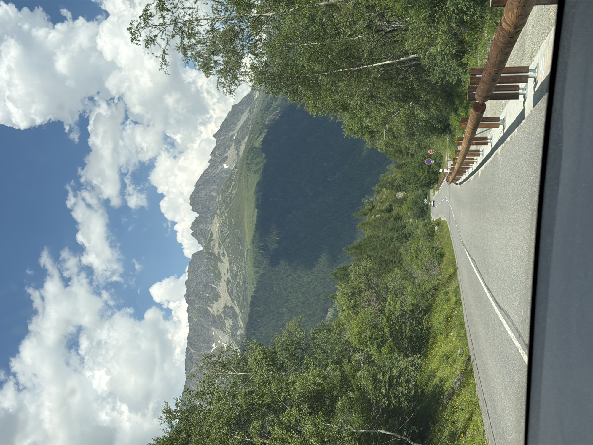 The image depicts a scenic mountain road in Vallorcine, France. The road curves gently upwards, bordered by a wooden guardrail and lush green trees on either side. In the distance, a majestic mountain range dominates the skyline, with its peaks partially obscured by dramatic clouds. The sky is a vibrant blue, interspersed with fluffy white cumulus clouds. A few cars are visible on the road in the distance, suggesting a mild level of traffic. The environment appears to be a natural, undeveloped area, characteristic of a mountainous region. The presence of a road and guardrail indicates it is a passable route for vehicles.