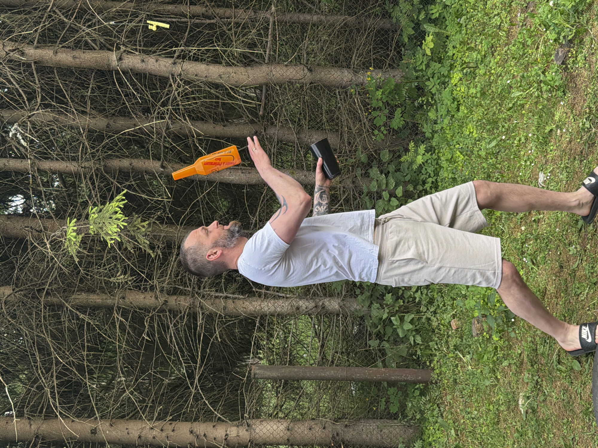 A man is shown in a side-profile view, mid-air toss of an orange bottle against a backdrop of dense coniferous trees. He is wearing a white t-shirt, light-colored shorts, and black flip-flops. The man has a beard and tattoos on his arms. He is holding a black cylindrical object in his left hand, preparing to catch the bottle. The environment is grassy with some foliage and a chain-link fence visible behind him. The scene appears to be outdoors, likely in a park or wooded area. There is no visible indication of the city of Tallinn, Estonia.