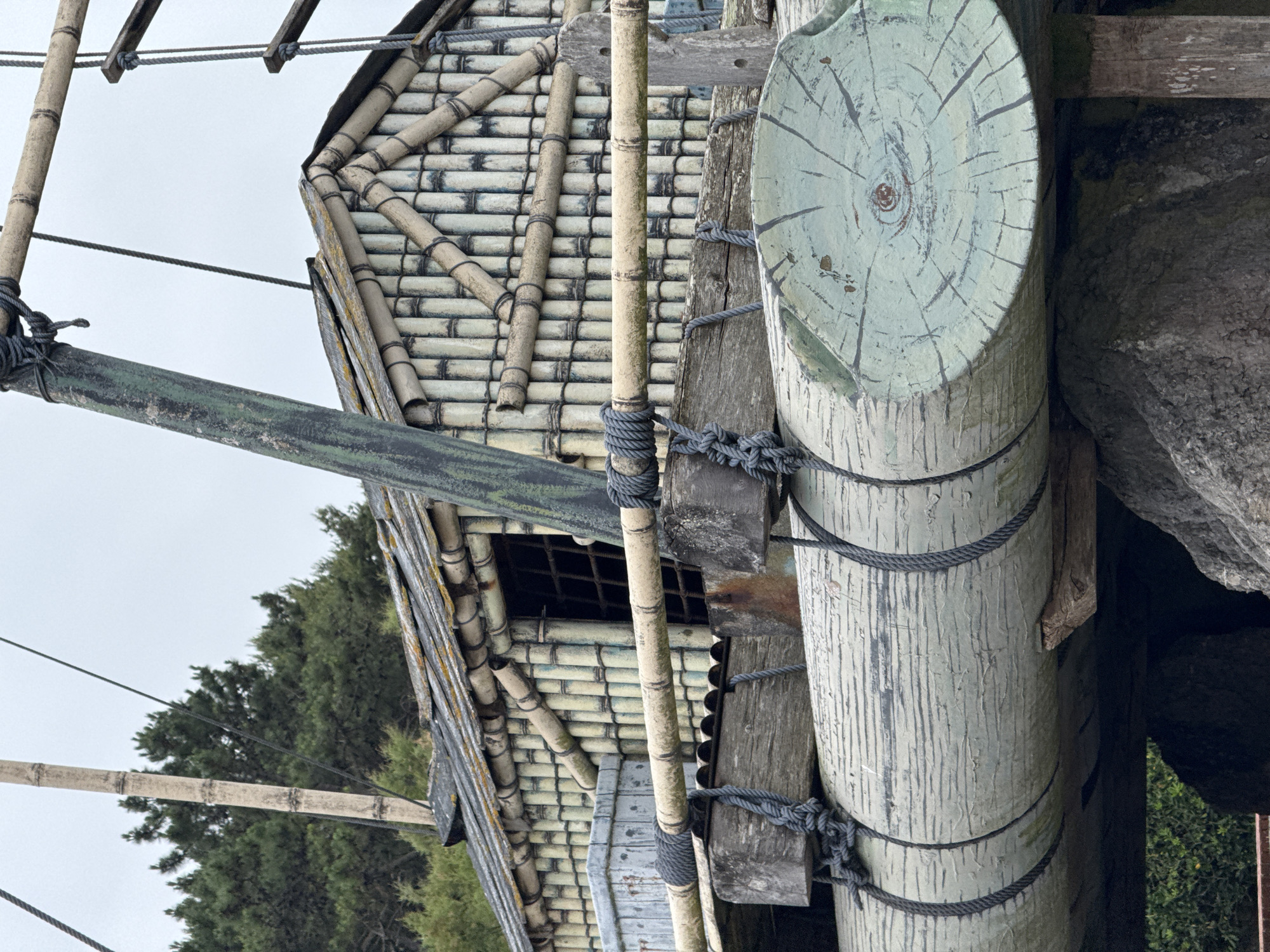 This image shows a close-up view of a raft-like structure constructed primarily from bamboo and logs. The raft supports a small, weathered structure resembling a hut or cabin, also built with bamboo. The roof of the hut appears to be made of slate or similar material. Ropes are used to lash the bamboo poles and wooden beams together. The raft itself is made of several large logs, painted with a greenish-blue hue and dark lines suggesting wood grain. One log end shows a painted design resembling a stylized eye. In the background, there is a dense line of green trees under an overcast sky. The raft is supported by large rocks and wooden posts, suggesting it is a permanent installation rather than a functional vessel. This scene is located in the Museum of Marine Cantabrica in the city of Santander, Spain, specifically the replica of the Kon-Tiki raft.