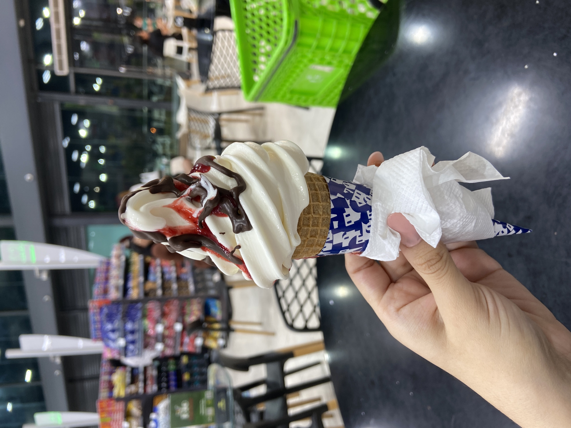 A person is holding a soft-serve ice cream cone with chocolate and strawberry syrup drizzled on top. The cone is wrapped in a blue and white paper wrapper and held with a tissue. The background shows a store interior with shelves of various products, tables, chairs, and a green shopping basket. The image may have been taken in or near Juan Griego, Venezuela.