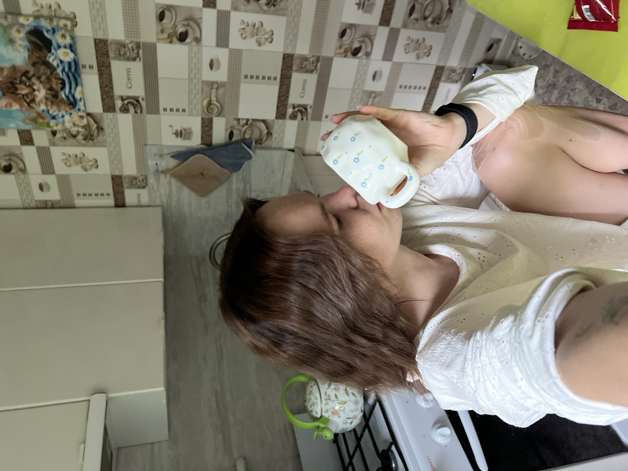 A woman is drinking from a white mug with a blue floral pattern in a kitchen. She is wearing a white blouse with lace details and a black watch on her left wrist. She has brown hair and fair skin. The kitchen has tile walls with a coffee-themed pattern. There is a kettle on the stove. The location is Chaikovsky, Russia.