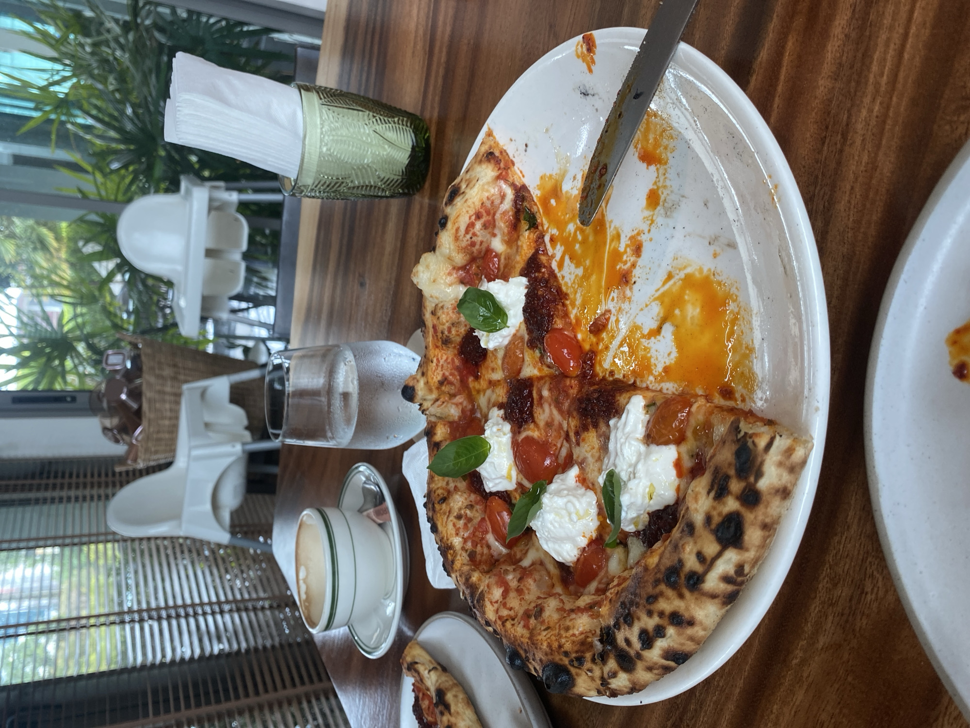 The image depicts a partially eaten pizza on a white plate, with a knife resting on the plate. The pizza is topped with tomatoes, white cheese, and basil. A glass of water and a cup of coffee are positioned near the pizza. A napkin holder and a high chair are visible in the background. The setting is a wooden table. The location is Choeng Thale, Thailand. No people are visible.