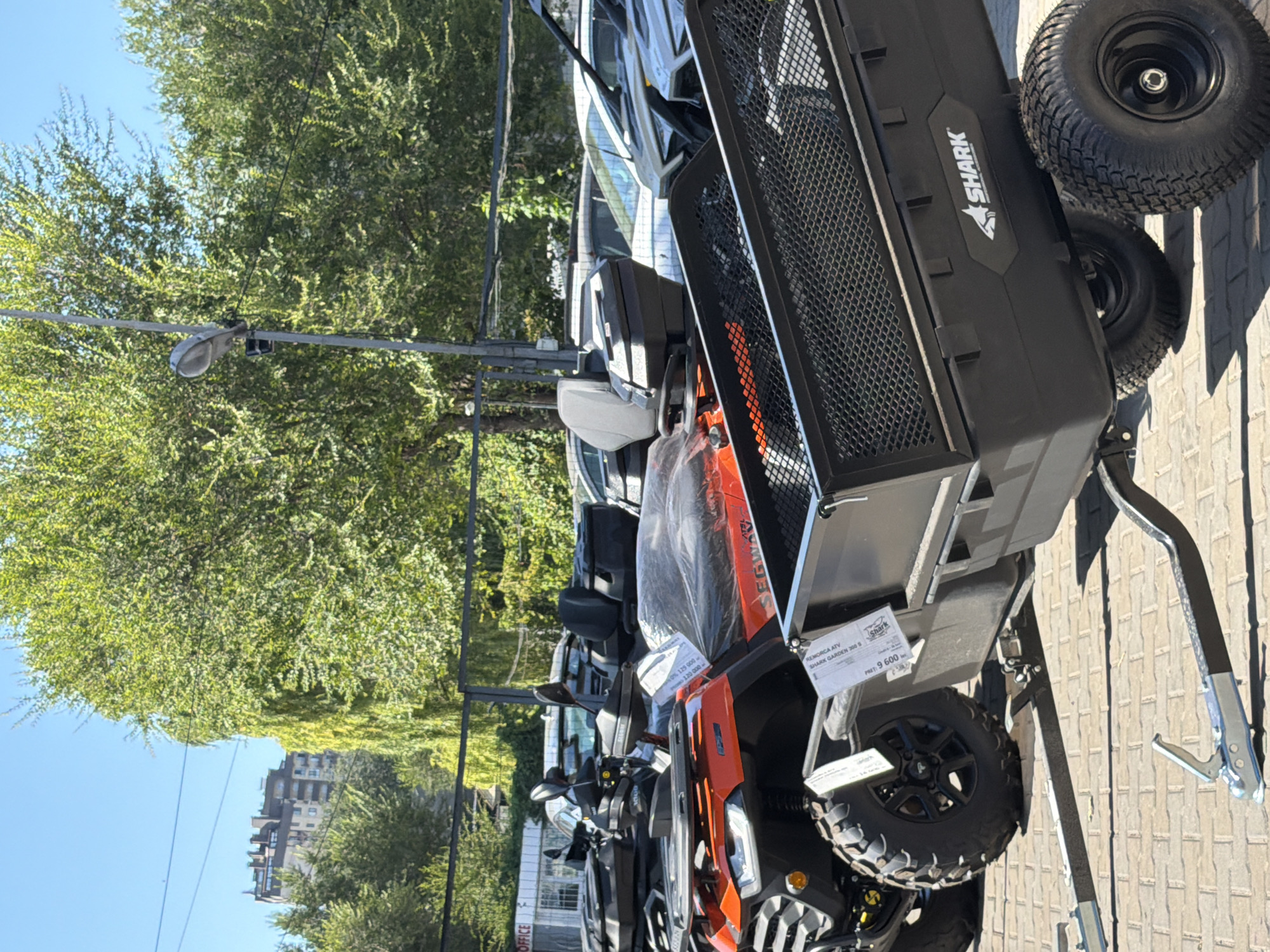 This is a close-up, eye-level, outdoor shot of several ATVs and a trailer parked on a paved surface. The ATVs are orange and black, with large, knobby tires. One ATV has a seat and handlebars visible. A black trailer with a mesh top is attached to the back of one of the ATVs. The trailer has "SHARK" logo on its side. In the background, there are trees, a building, and what appears to be a parking lot with other vehicles. The sky is clear and blue. The image does not contain any people. The scene is likely a dealership or an outdoor storage area for ATVs. The location in Chișinău, Moldova, is not explicitly indicated within the image itself.