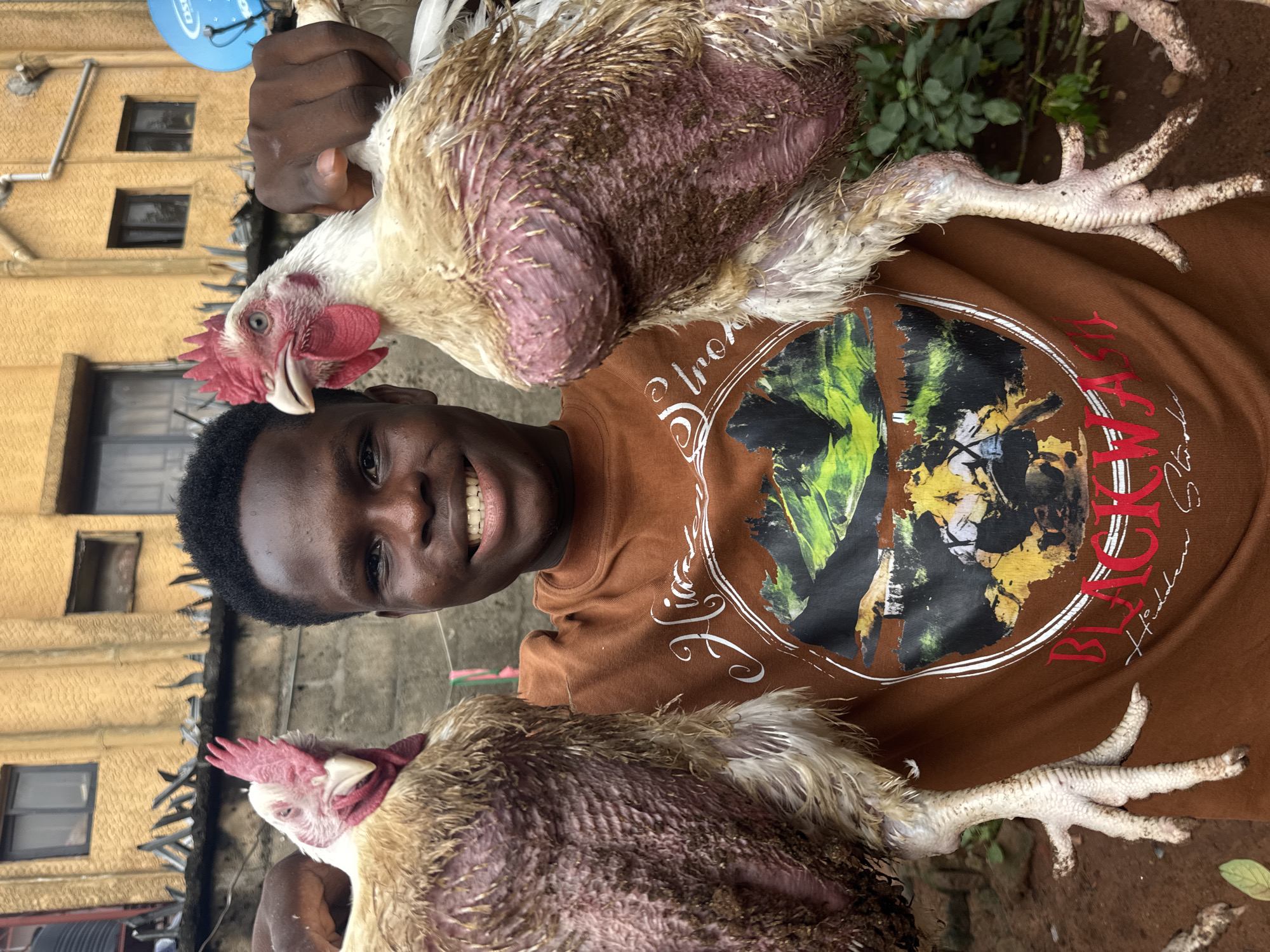 A young man with dark skin and short, curly hair is smiling broadly at the camera while holding two live chickens upside down, one in each hand. He is wearing a brown T-shirt with a large, elaborate graphic print on the front.
The chickens have white and light brown feathers, with prominent red combs and wattles. Their bodies, particularly their undersides, appear to have sparse feathering, revealing pinkish-purple skin with visible feather shafts and some dirt adhering to the skin and remaining feathers. The man is holding each chicken by its legs.
In the background, there are yellow-painted concrete or stucco buildings with dark-framed windows, suggesting an urban or semi-urban residential setting, consistent with Onitsha, Nigeria. Part of a blue satellite dish, clearly branded "DStv," is visible attached to one of the buildings. Green foliage can be seen near the bottom of the frame, and the ground appears to be dirt or concrete. The lighting suggests it is daytime.
Visible text on the man's T-shirt includes the word "BLACK" in a stylized font, with other fragmented words like "BLACKWΛ" or "BLΛCK HΛLL" and some illegible script.