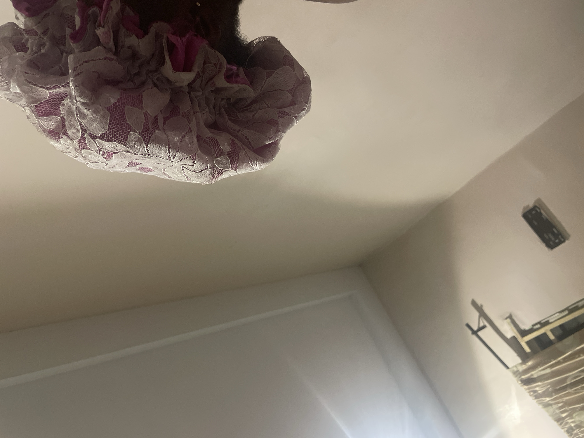 A close-up, low-angle shot shows the top of a person's head wearing a pink and white lace bonnet. The bonnet is gathered at the top and has a floral lace pattern over a pink fabric. The background is a plain, light-colored wall and ceiling, with a section of a curtain visible in the lower left corner. The overall lighting is dim. The image was taken in Jalingo, Nigeria.