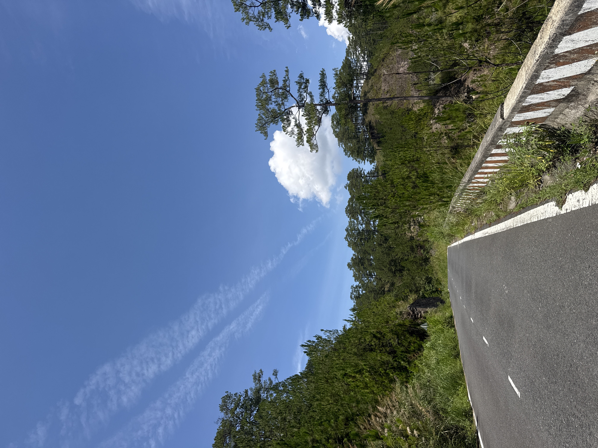 The image depicts a roadway in Hiep Thanh, Vietnam, bordered by a guardrail and lush vegetation, with a clear blue sky overhead featuring contrails and a single cumulus cloud. The asphalt road is marked with a white line, indicating lane division. The guardrail shows alternating light and dark stripes. Abundant green trees and grassy growth flank the roadway. No people are visible.