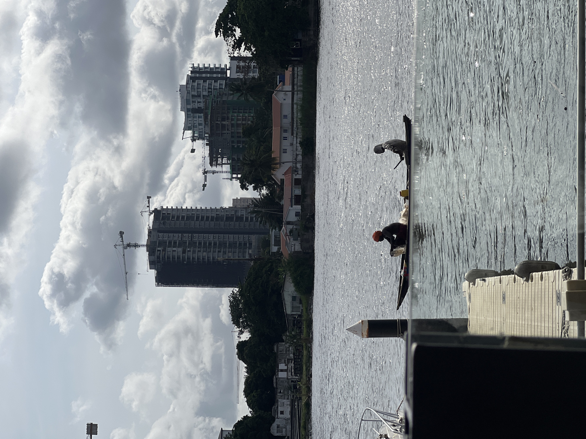 The image shows a waterfront scene in Ikoyi, Nigeria, with construction cranes towering over buildings along the shoreline. Two individuals are on stand-up paddleboards in the water. One is wearing a helmet and dark clothing, while the other is wearing lighter clothing. The water surface reflects light, and a marina or dock structure is visible in the foreground. Clouds dominate the sky.