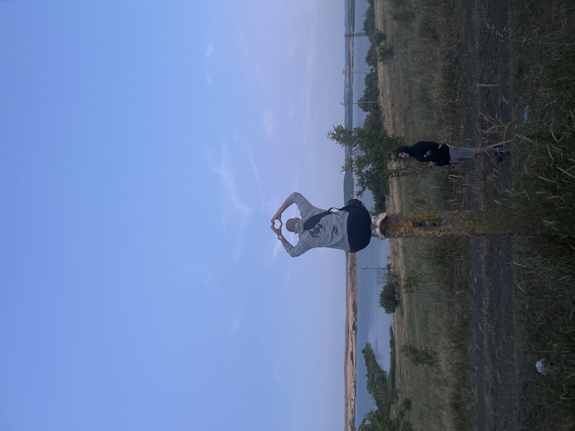 Two individuals are present in an outdoor scene with a body of water in the background. A person is seated atop a vertical, rough-textured post, facing away from the viewer, and has formed a heart shape with their hands above their head. This individual is wearing a gray hoodie with a black backpack and dark shorts. To the right and slightly behind, a second person wearing a black hooded sweatshirt and gray shorts stands on the ground, holding what appears to be a phone. The environment consists of dry grass, scattered bushes, and a distant view of hills and a body of water, likely a lake or bay. The sky is a pale blue with light clouds, indicating it is either dawn or dusk. The overall setting suggests a rural or semi-natural area.