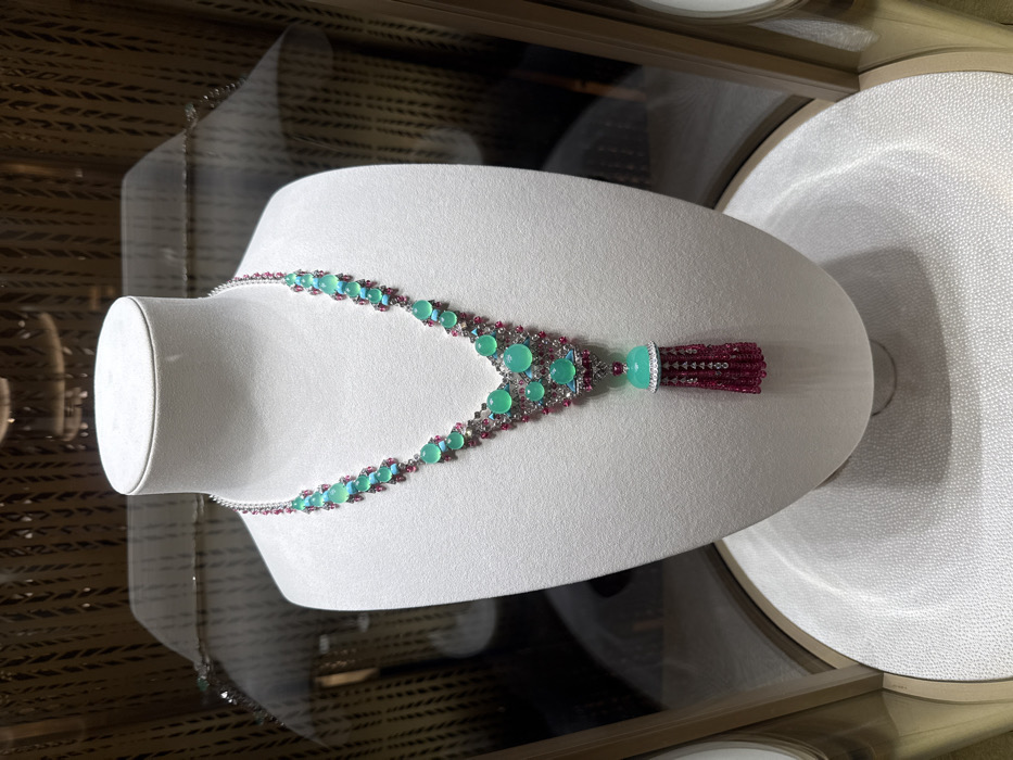 A mannequin neck displays a necklace with alternating green and red stones. Smaller red stones connect larger green ones. The necklace culminates in a pendant with a larger green stone and a red tassel. The display is set inside a glass case with reflective surfaces visible in the background. A circular white textured platform is to the right of the mannequin.