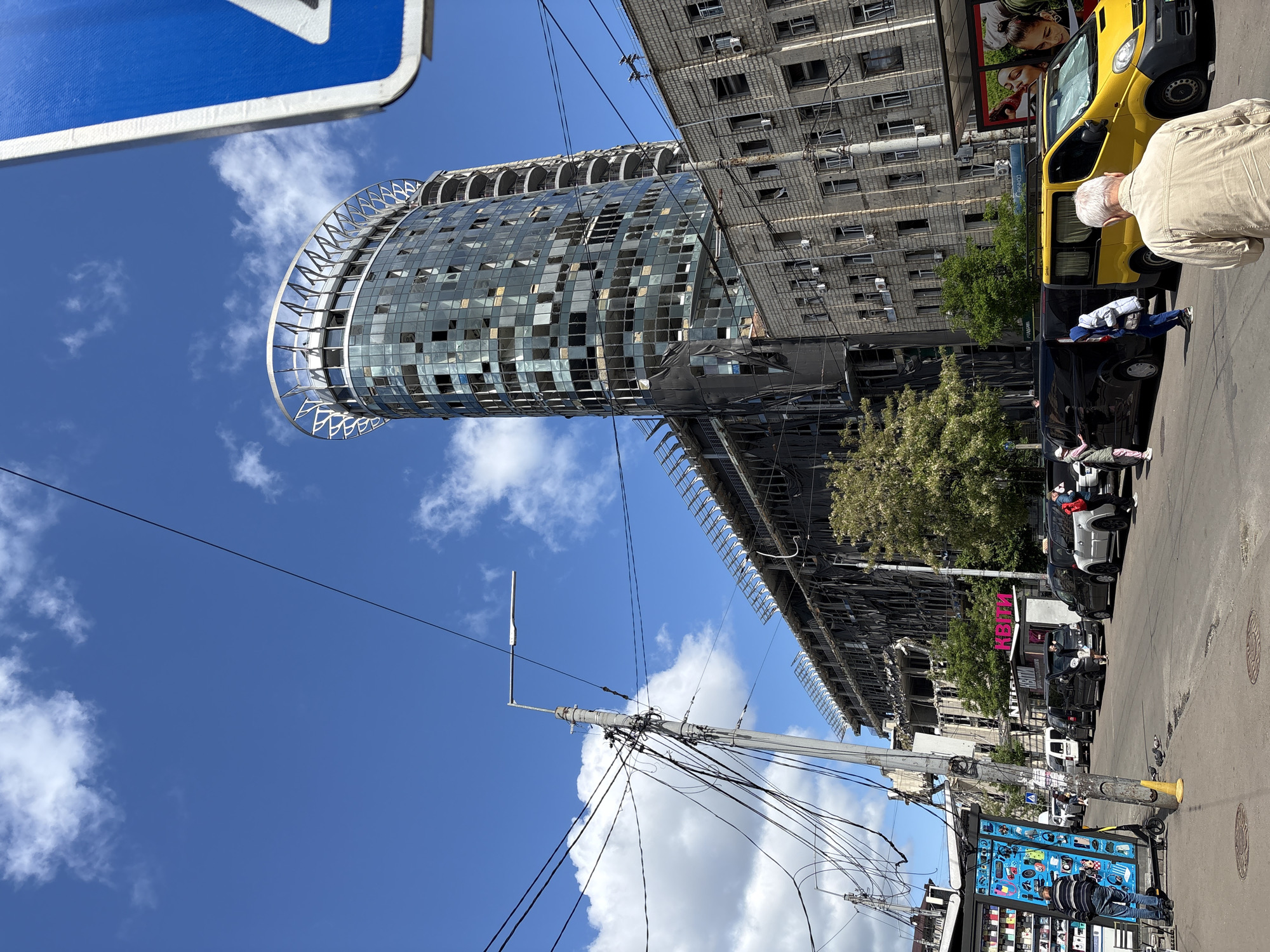 The image shows a street scene in Kyiv, Ukraine. The most prominent feature is a severely damaged, partially collapsed, cylindrical-shaped building with a steel skeletal structure and shattered glass. Next to it, a traditional Soviet-era apartment block displays visible damage and signs of destruction.
People are present on the street, including a man with grey hair standing near a yellow van. Several dark-colored cars are parked along the roadside. A commercial sign with the Cyrillic word "КВІТИ" (Kvitky) meaning "Flowers" is visible. A utility pole with numerous overhead wires is also featured in the foreground.
The sky is blue with scattered white clouds. A blue street sign is partially visible in the upper-left corner.