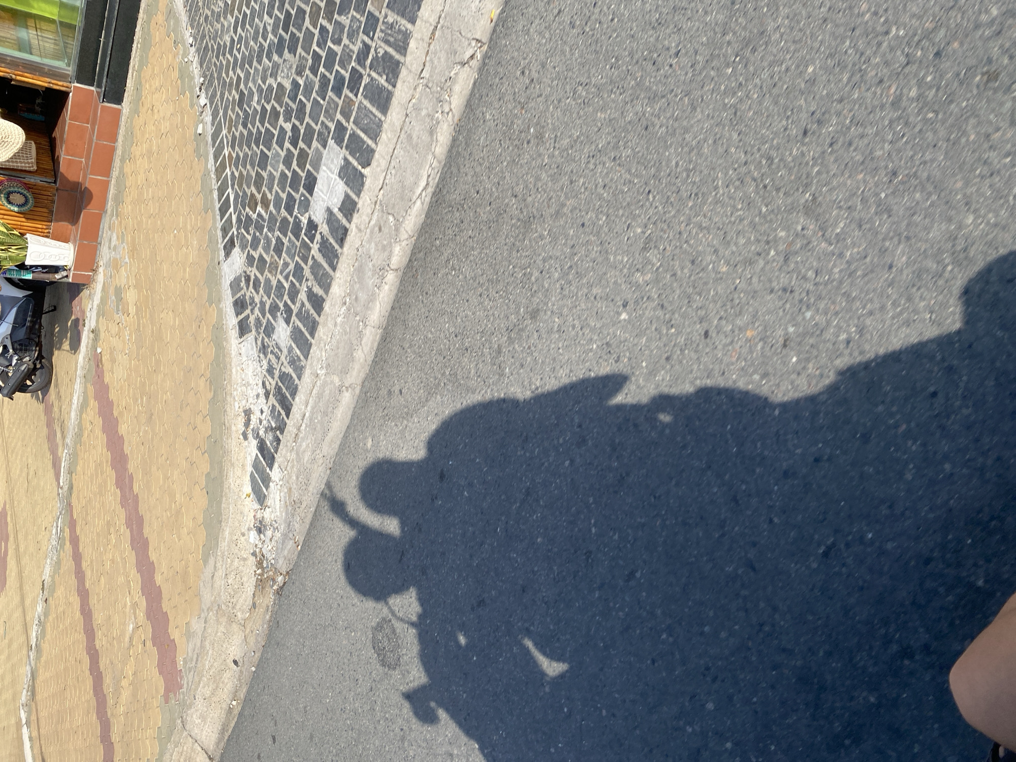 In Phu Thuy Ward, Vietnam, a long, dark shadow of two people on a motorcycle stretches across an asphalt road. The shadow shows two figures, one presumably the driver with hands on the handlebars, and another passenger behind. The motorcycle itself is partially visible in the background as a dark shape with a wheel. The road is bordered by a textured sidewalk made of interlocking yellow and red pavers. Beyond the pavers, a darker, brick-like section of the sidewalk is visible. The scene is illuminated by bright sunlight, casting sharp shadows and highlighting the textures of the road and sidewalk. The overall impression is of a typical street scene in Vietnam on a sunny day.