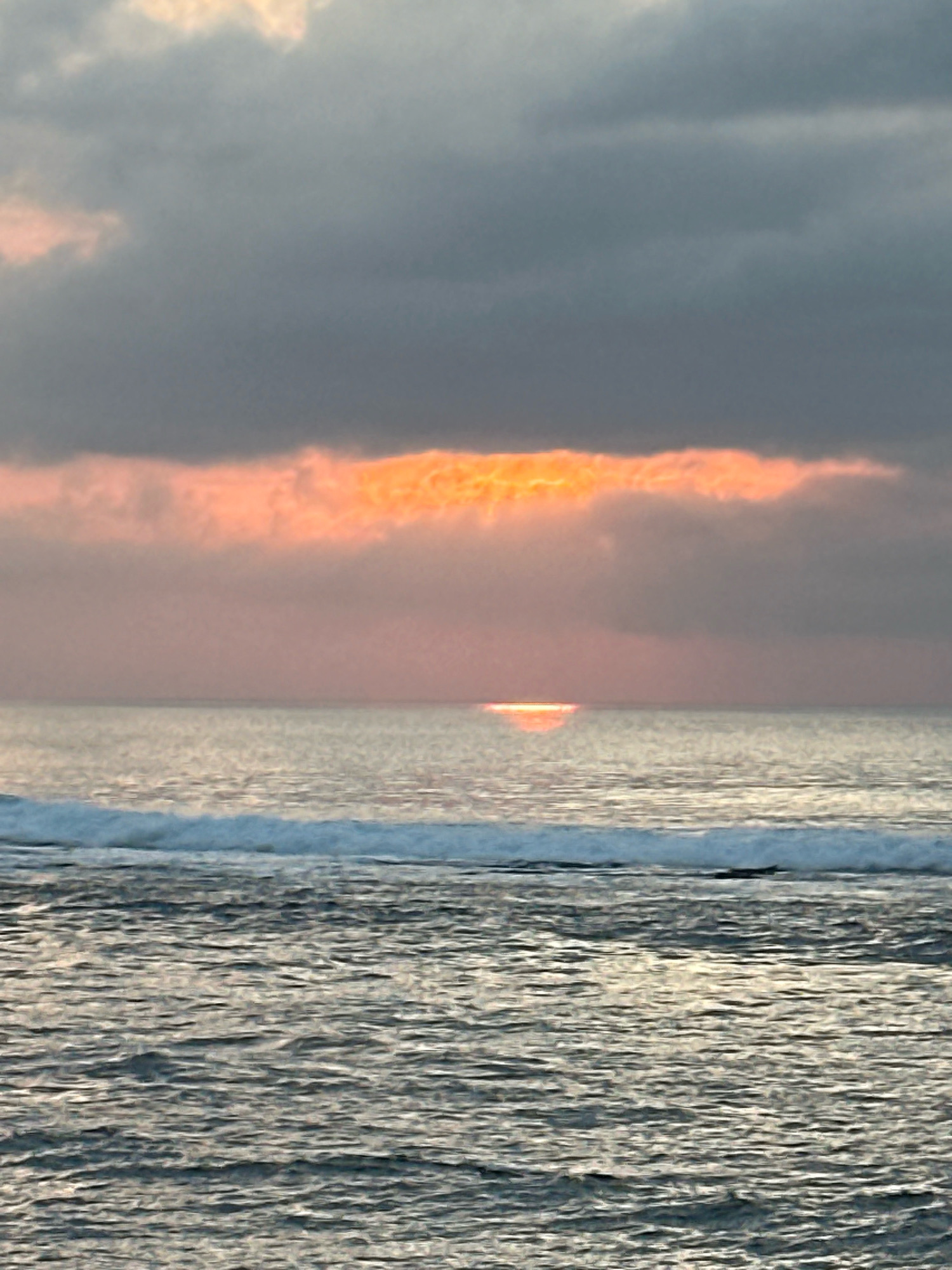 A serene seascape is depicted at sunset in Blahbatuh, Indonesia. The sky is filled with dramatic, heavy gray clouds, but a horizontal band of vibrant orange and pink light pierces through the clouds, casting a warm glow. The sun, a bright, luminous orb, is partially obscured by the clouds, its reflection shimmering on the calm sea. The water itself is a deep, tranquil blue-grey, with subtle ripples and the gentle crest of a wave in the foreground, its white foam contrasting with the darker water. The overall scene is one of peaceful, natural beauty, characterized by the interplay of light and shadow on the water and in the sky.