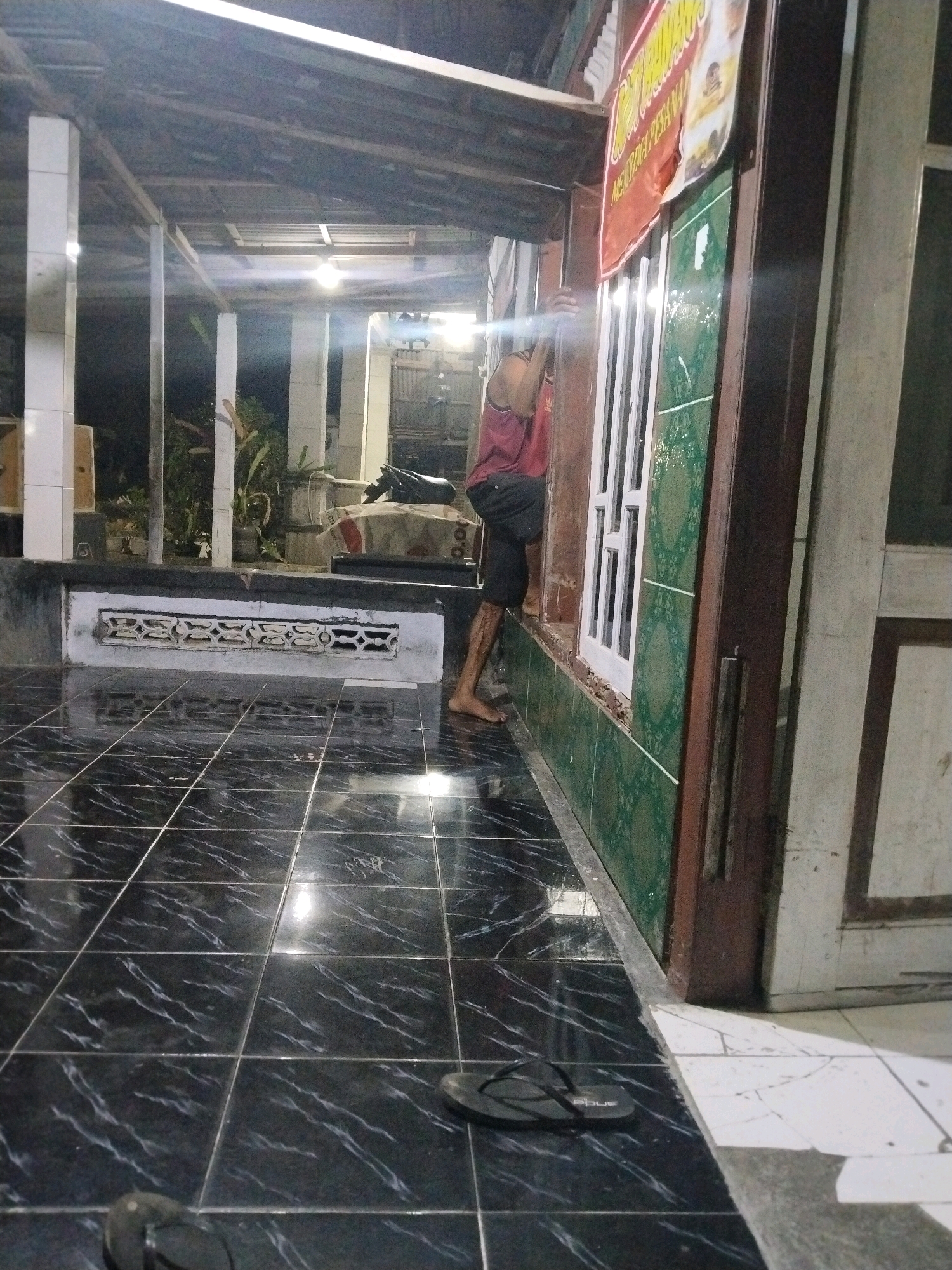 A person is observed climbing into a building through a window. The scene appears to be an outdoor area, possibly a shop or residential building, at night. The person is wearing a red tank top and black shorts. The building has a tiled facade with green patterned tiles and a wooden frame around the window. The ground is covered in shiny black tiles. A sign with red and yellow lettering is partially visible above the window. In the background, there are pillars, some plants, and what looks like a motorcycle parked. The lighting suggests artificial light sources are present. The location is identified as Pandan Blole, Indonesia.