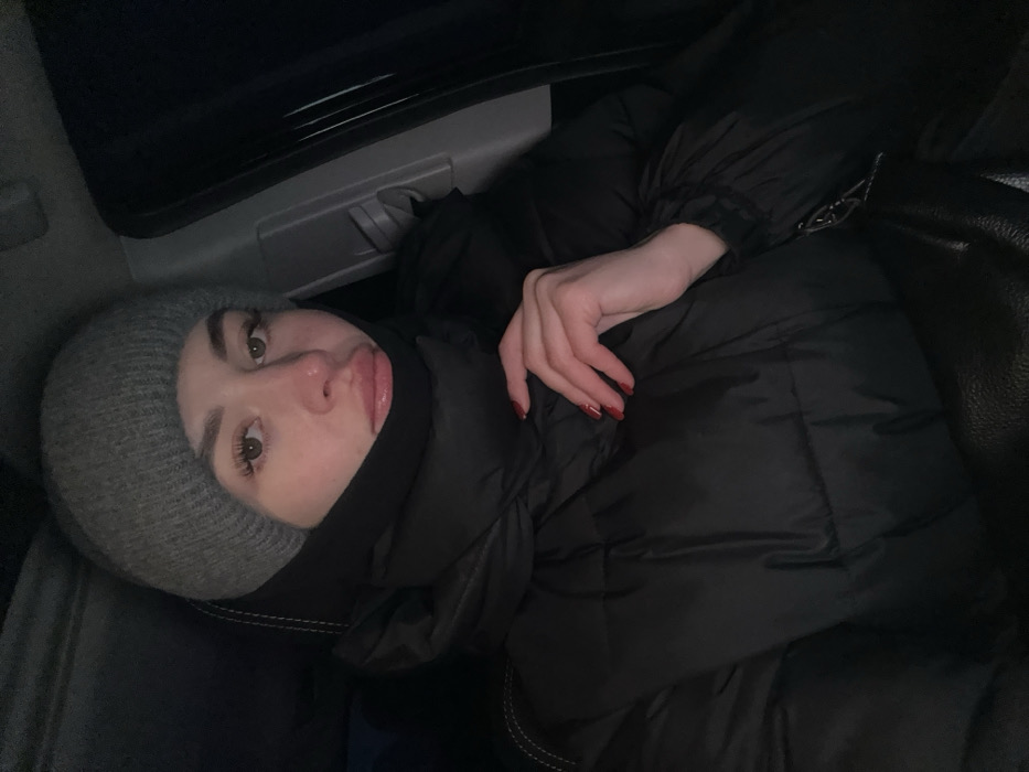 A person in a gray knit hat and black face covering is lying down in what appears to be a car or dark interior space. They are wearing a black puffer jacket and their pale hand, adorned with red nail polish, is resting on the jacket. The individual is looking at the camera with a slightly parted mouth, appearing to be pouting. The lighting is dim, casting shadows around the scene. A car door handle is visible in the background.