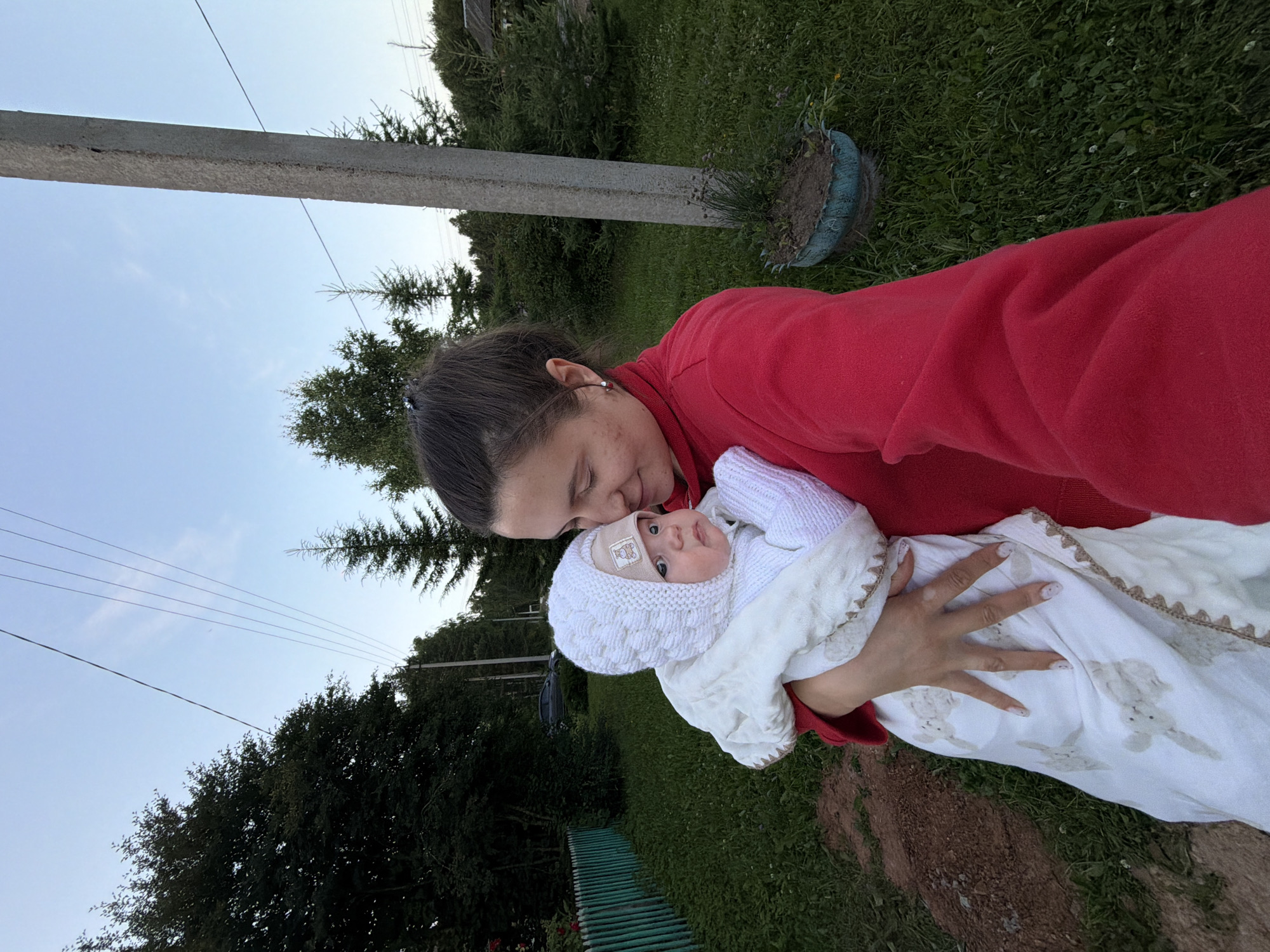 In Красный Маяк, Russia, a woman in a red sweater holds a baby wrapped in a white blanket and wearing a white knitted hat. The baby looks towards the camera with a neutral expression. The background features a grassy area, a green wooden fence, a concrete pole, and trees, indicating a rural or semi-rural setting. The sky is clear and blue, with power lines visible overhead. A car is parked in the distance. The overall scene captures a moment of tenderness between a mother and her child outdoors.