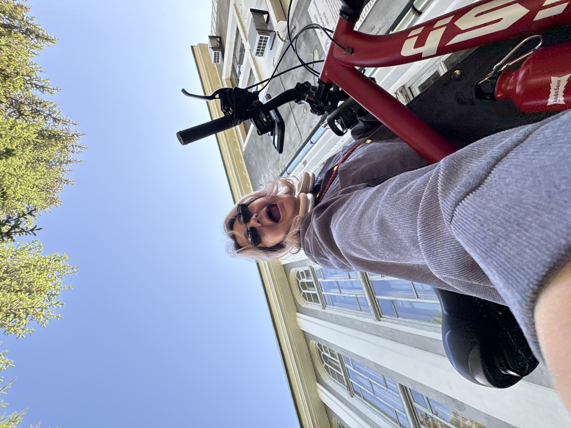 A low-angle, outdoor, wide shot shows a person on a red bicycle in front of a light-colored building with tall, arched windows and green trees overhead. The person, with blonde hair and wearing sunglasses and a grey jacket, is in the foreground, looking at the camera with their mouth open. The bicycle's handlebars and a portion of the frame are visible. A red water bottle is attached to the bicycle. The building appears to be located in Birobidzhan, Russia.