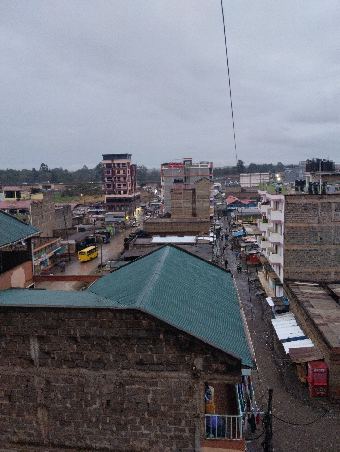 An overcast day in a densely populated urban area shows buildings of varying heights and materials. A yellow bus moves along a wet street lined with shops. Close-up buildings in the foreground are made of brick and have green roofs. A single cable stretches across the sky. Laundry hangs on a balcony. The image depicts the scene from an elevated viewpoint, overlooking the urban sprawl.