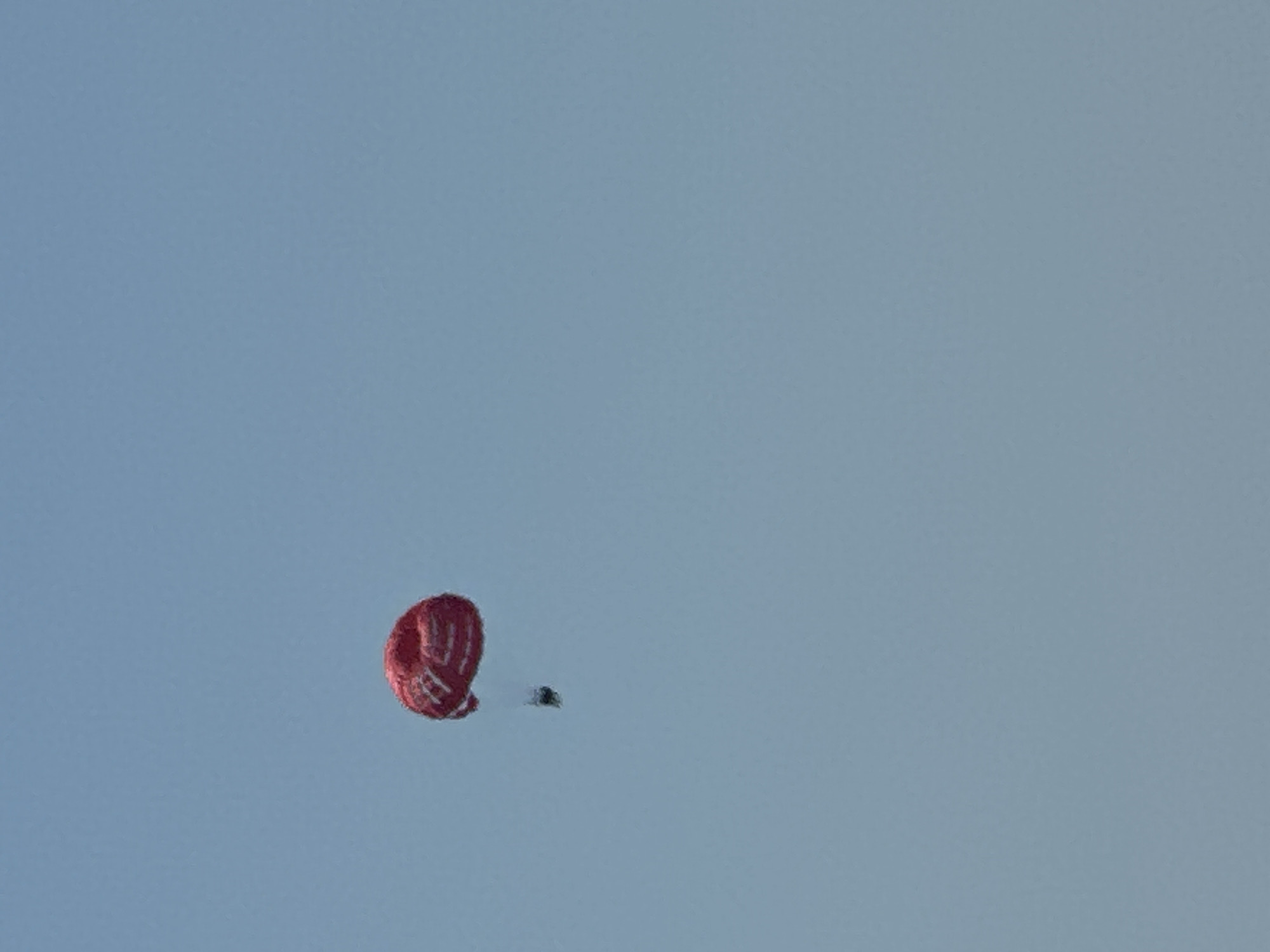 A single person is parasailing in the sky above Kamenovo, Montenegro. The parasail is red with white stripes. The sky is clear blue, indicating fair weather. The image captures the person mid-air, suspended by the parasail.