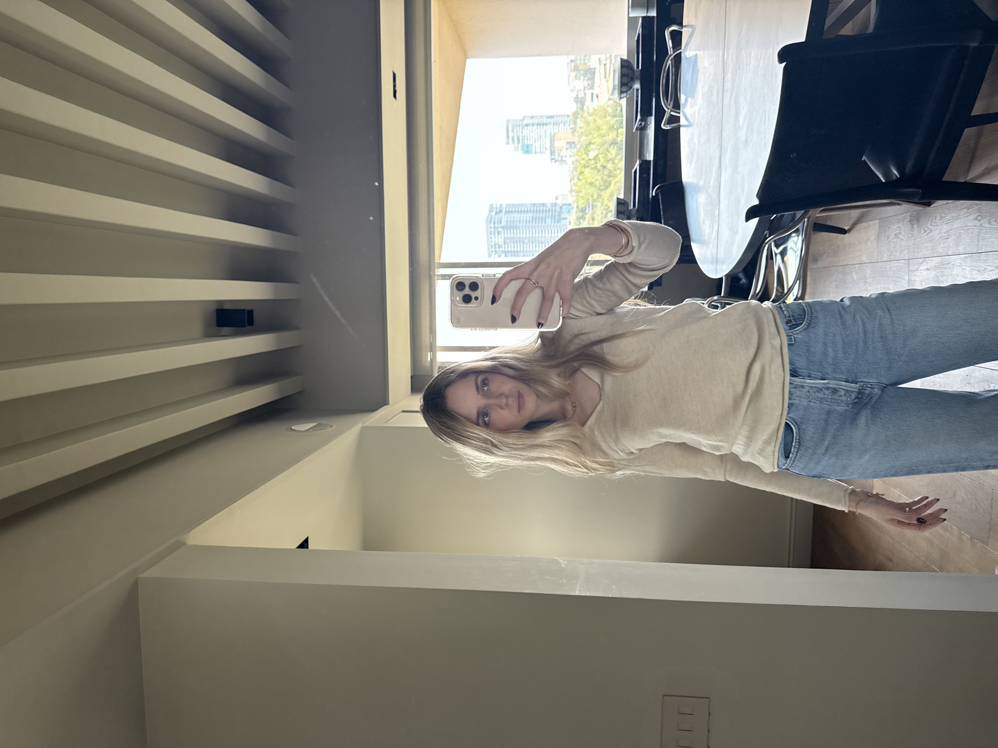 A young woman is taking a selfie in what appears to be an apartment or condo in Mexico City, Mexico. She is wearing light blue jeans and a beige long-sleeved shirt. The background shows a view of the city with tall buildings. A dining table and chairs are visible to the right of the frame. The apartment appears to have a modern interior design with a unique ceiling feature.
