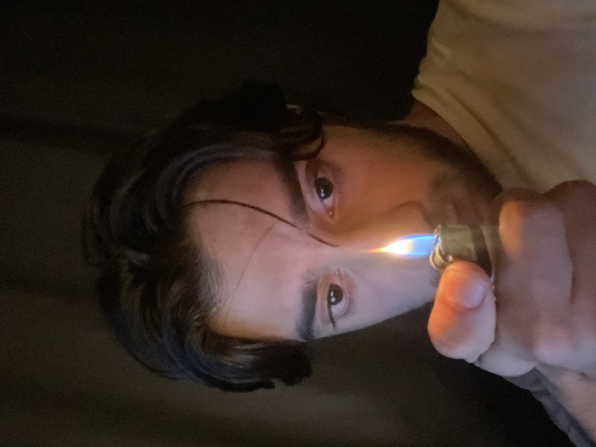 A close-up, low-angle, portrait of a young man in Huixquilucan de Degollado, Mexico, holds a lit lighter towards the camera. The man has dark, wavy hair that falls across his forehead and is looking intently at the flame. His right eye is visible, reflecting the light from the lighter. His skin is illuminated by the warm glow of the flame, creating a dramatic contrast with the dark background. He is wearing a light-colored t-shirt. The lighter is metallic and positioned in his right hand, with the flame a bright orange and blue. The overall ambiance is intimate and dramatic, with the focus sharply on the man's face and the lighter.