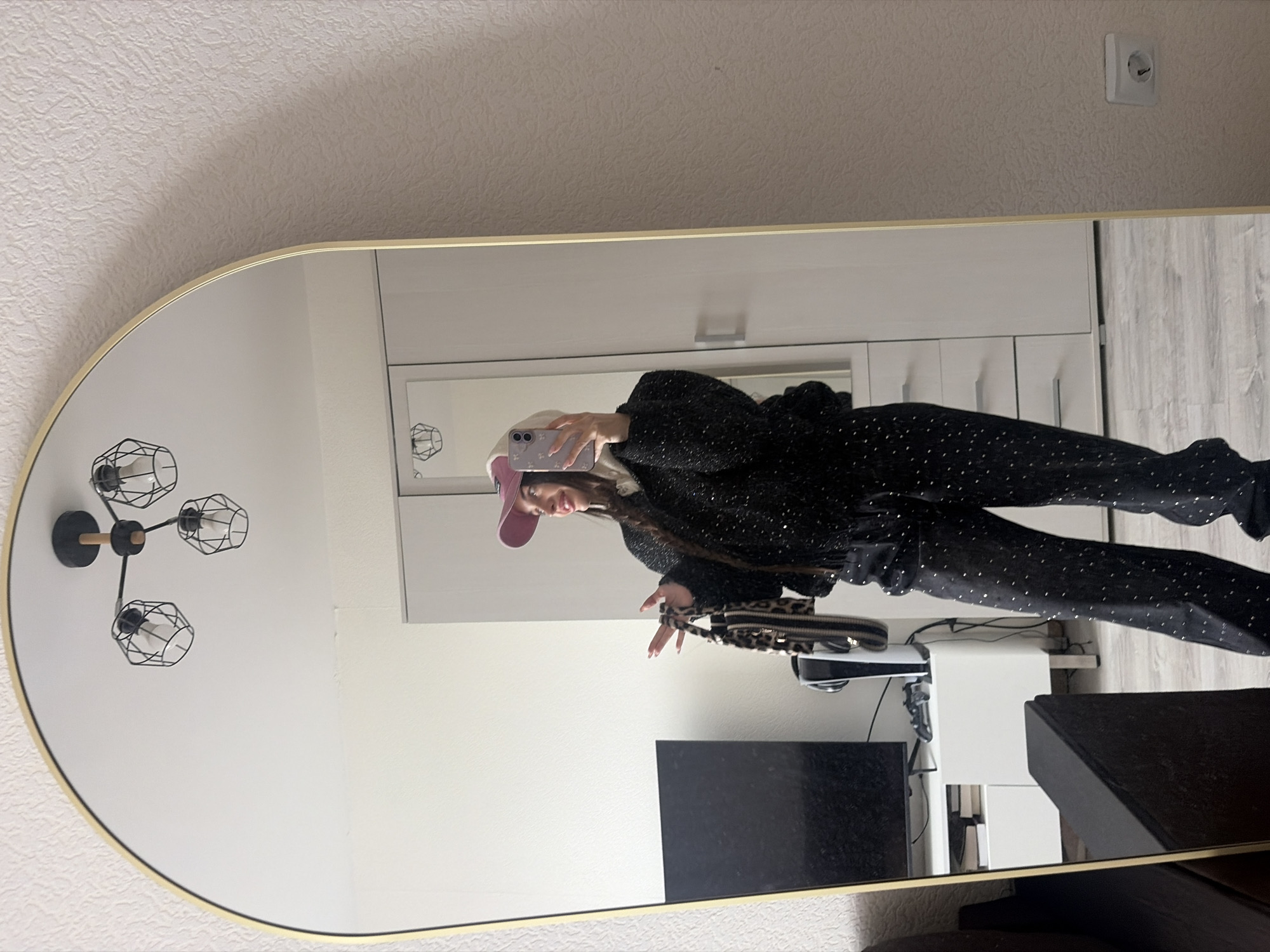 The image shows a person reflected in a mirror, likely in an indoor setting. The person is wearing a black outfit with what appears to be silver embellishments, a pink baseball cap, and has a long braid. They are holding a mobile phone, apparently taking a selfie in the mirror. A patterned handbag or purse is visible. The background includes a white dresser or storage unit, a wall-mounted light fixture with a geometric design, a TV, and other furniture items. No location information regarding Saint Petersburg, Russia, is discernible from the image.