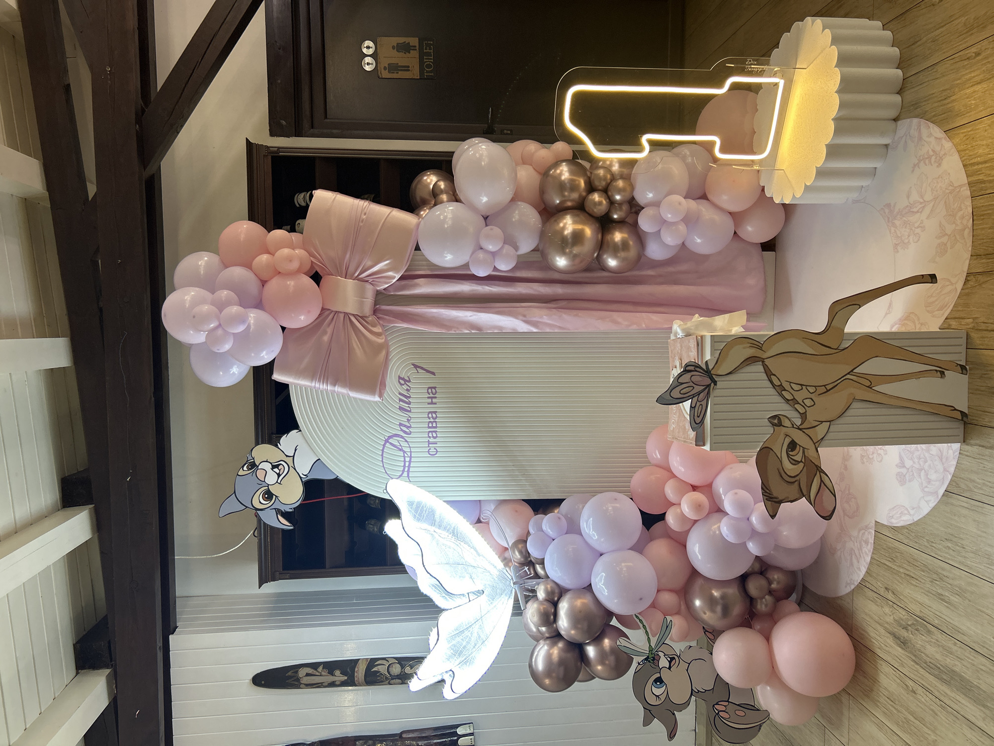 The image depicts a birthday party decoration, likely celebrating a child's first birthday. The color scheme is predominantly pastel pink, white, and rose gold. A large, stylized number "1" is illuminated with neon lighting. Decorations include balloon garlands in various shades of pink, white, and rose gold, arranged around a decorative backdrop panel with the name "Далия става на 1" written in Cyrillic, meaning "Dalia is turning 1." Cardboard cutouts of Bambi and Thumper are incorporated into the decoration. There is a large, illuminated butterfly decoration as well. The setting appears to be indoors, with wooden beams and paneling visible, suggesting it might be a restaurant or event space. The presence of a toilet sign with male and female figures indicates a public venue. It is impossible to confirm if this is in Plovdiv, Bulgaria based only on this image.
