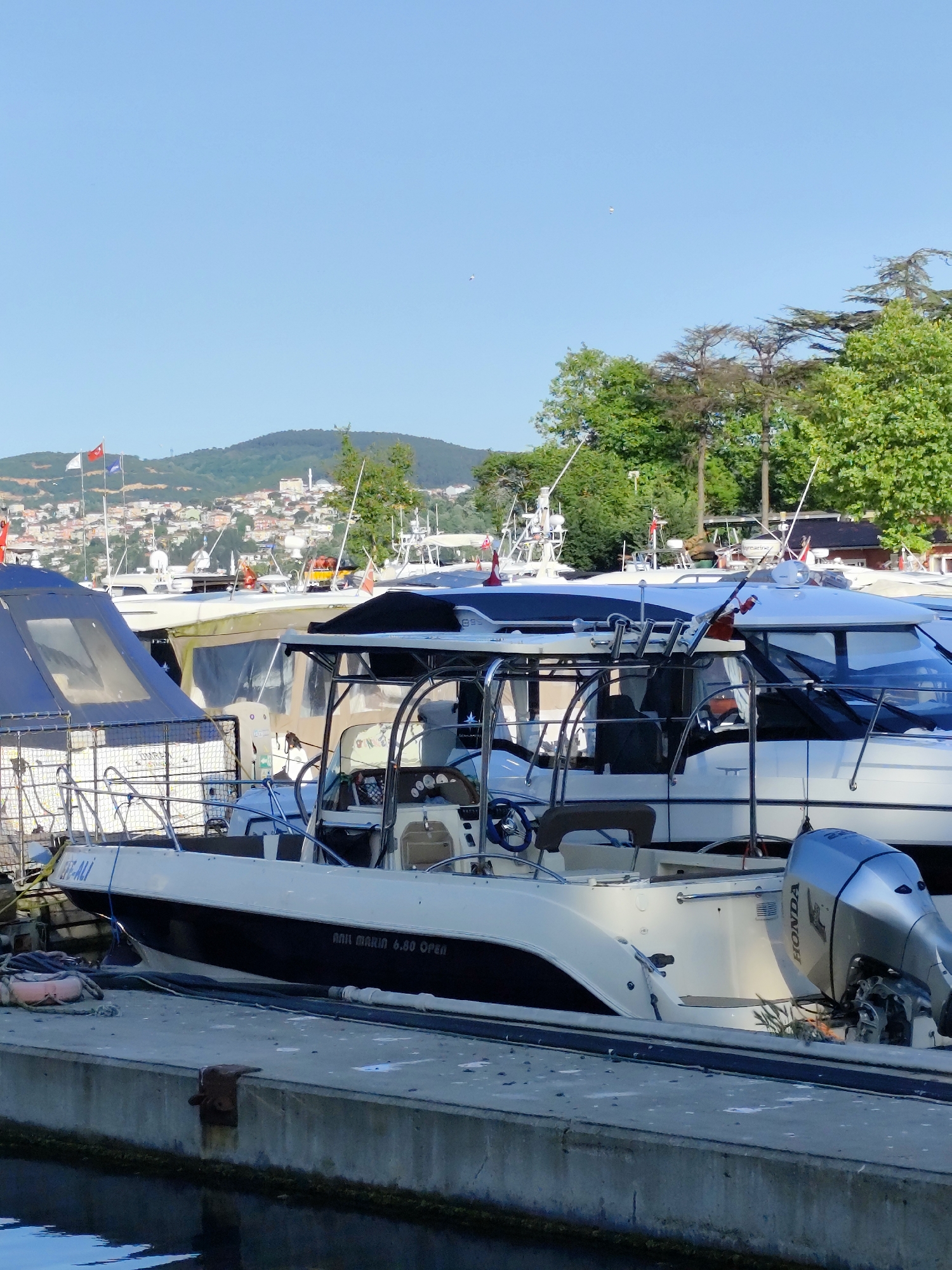 The image depicts a marina with several boats docked along a concrete pier. The foreground features a white and black boat with a Honda outboard motor. The boat is labeled "ARIL MARIN 6.60 OPEN." Other boats of varying sizes and designs are docked behind it, creating a layered view. In the background, a cityscape sprawls across a hillside, suggesting an urban setting with a mix of buildings and vegetation. Flags are visible atop some of the buildings. The presence of trees in the background suggests proximity to a green area. Based on the description of the city, the image seems to be in Sarıyer, Turkey. The sky is clear and blue, indicating a sunny day. The boats are motionless, suggesting they are currently at rest. The pier shows signs of wear and tear, with some algae growth visible along its edge.