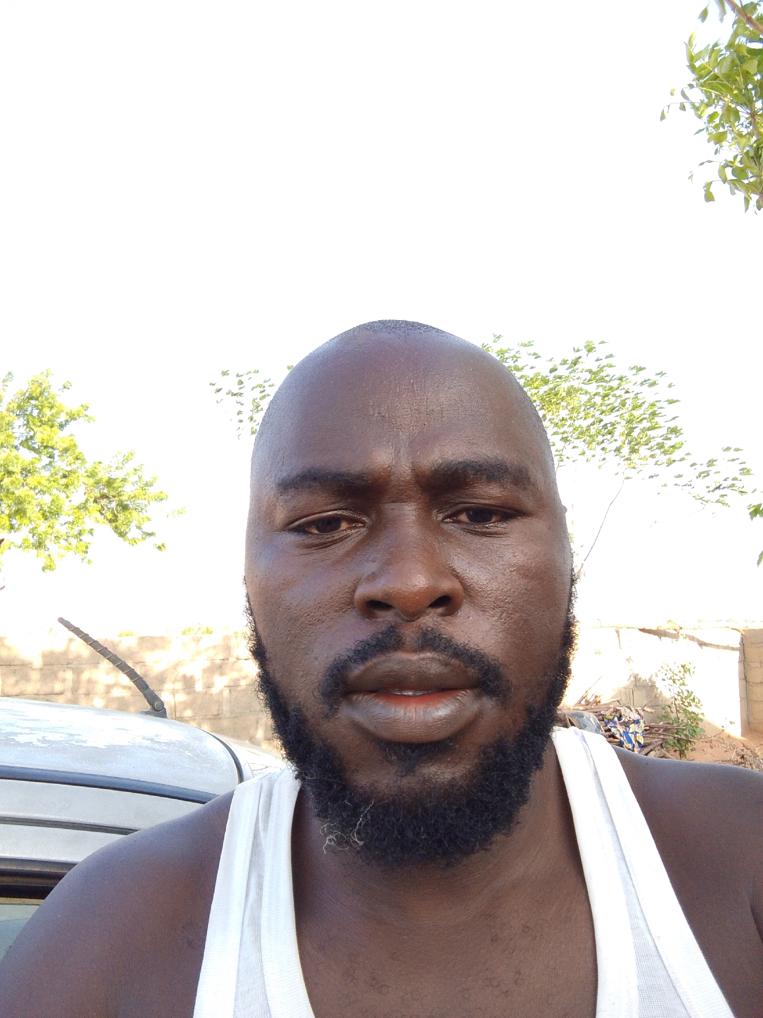 A close-up selfie of a dark-skinned man with a shaved head and a full beard. He is wearing a white tank top. In the background, there is a light-colored car with its antenna visible, sparse trees, and a rudimentary building, suggesting a rural area within Mulke Barikin Sayejo, Nigeria. The light is bright and suggests it is daytime. The man's expression is neutral.