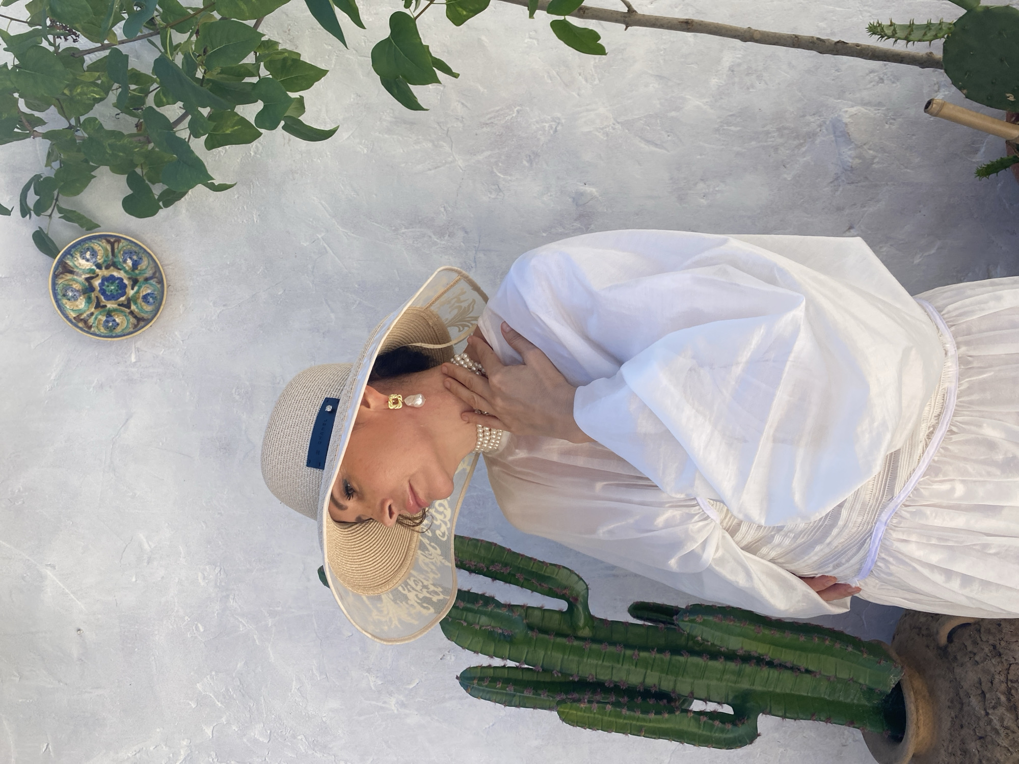 A woman wearing a wide-brimmed straw hat and a white flowing dress is posed outdoors against a textured gray wall. Her hand is resting on her neck, and she is adorned with pearl jewelry and large square earrings. A green cactus plant in a pot is positioned in the foreground. In the upper left corner, a decorative blue and green ceramic plate is visible, and overhead, green foliage hangs down. A bamboo-like object is seen to the right of the cactus. The scene appears to be set during the daytime, with soft lighting. The woman's gaze is directed away from the camera. A label with the text "GRAPEL" is visible on the side of her hat. The overall impression is one of elegant leisure.