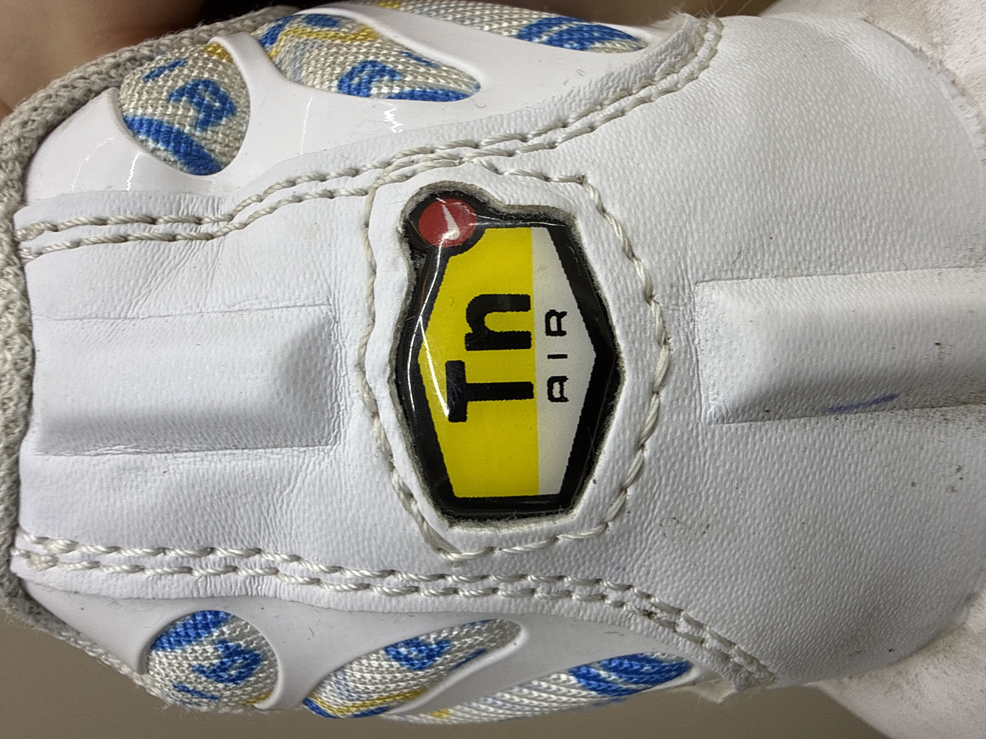 The image is a close-up, high-angle shot of a white Nike Air Max Plus TN sneaker, likely a child's size due to the prominent branding. The sneaker is the sole subject, captured against a neutral background that hints at a soft, possibly fabric surface. The material of the sneaker appears to be a combination of white leather with textured detailing and woven fabric panels featuring a blue, yellow, and white geometric pattern. Prominently displayed on the side of the shoe is the iconic "Tn Air" logo within a black, glossy rectangular frame. The "Tn" is depicted in bold black lettering, with the "T" being partially obscured by a small red circle with a white Nike swoosh. The "Air" is written in smaller black text to the right of the "Tn". The stitching on the shoe is a light beige, creating a visible pattern around the various panels. The overall condition of the shoe suggests it has been worn, with some minor scuffs and dust visible on the white surfaces. The lighting is bright, emphasizing the textures and colors of the sneaker. No people, specific setting details beyond the sneaker itself, time of day, or weather are discernible. The only visible text is the "Tn Air" branding. The context of Burgas, Bulgaria, does not provide any direct visual cues within the image.