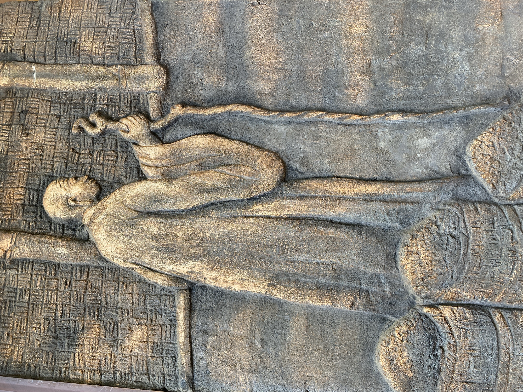 A low-relief sculpture is set into a gray, textured background. The sculpture depicts a reclining figure in profile, facing left. The figure is wearing a long, flowing garment with the head of the individual facing towards the top left of the image.
The figure holds an object with a curved end, the fingers grasping it. A twisted rope-like element extends from the figure's hand, continuing along the upper part of the sculpture. To the right of the figure, there are round shapes, possibly barrels, with textured details suggesting wooden construction. Behind the figure, to the left, is a textured area with a pattern reminiscent of woven material.
This sculpture is located at Domaine de Marie Church in Da Lat, Vietnam.