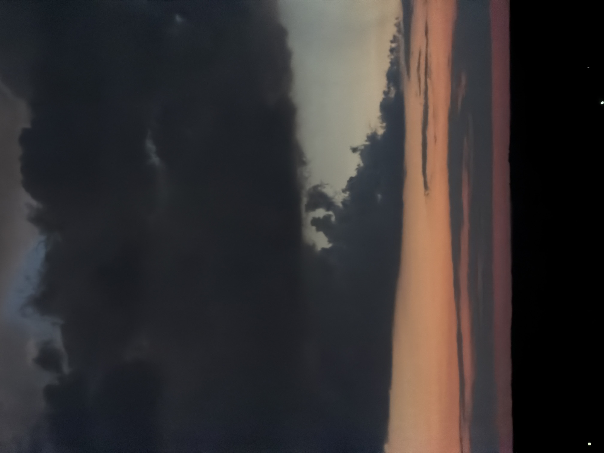 The image depicts a dark, ominous sky with heavy, bruised clouds dominating the upper two-thirds of the frame. Below the clouds, a band of sunset colors, ranging from pale orange to soft pink and gray, is visible. The lower portion of the image is entirely black, obscuring any ground detail, though two small white specks are visible in the bottom left and right corners, possibly indicating distant lights or reflections. The overall impression is one of an approaching storm or twilight. The location is identified as Unknown, Unknown. There are no people, subjects, or specific objects clearly discernible in the scene, only the atmospheric elements of the sky and a silhouette of the ground.