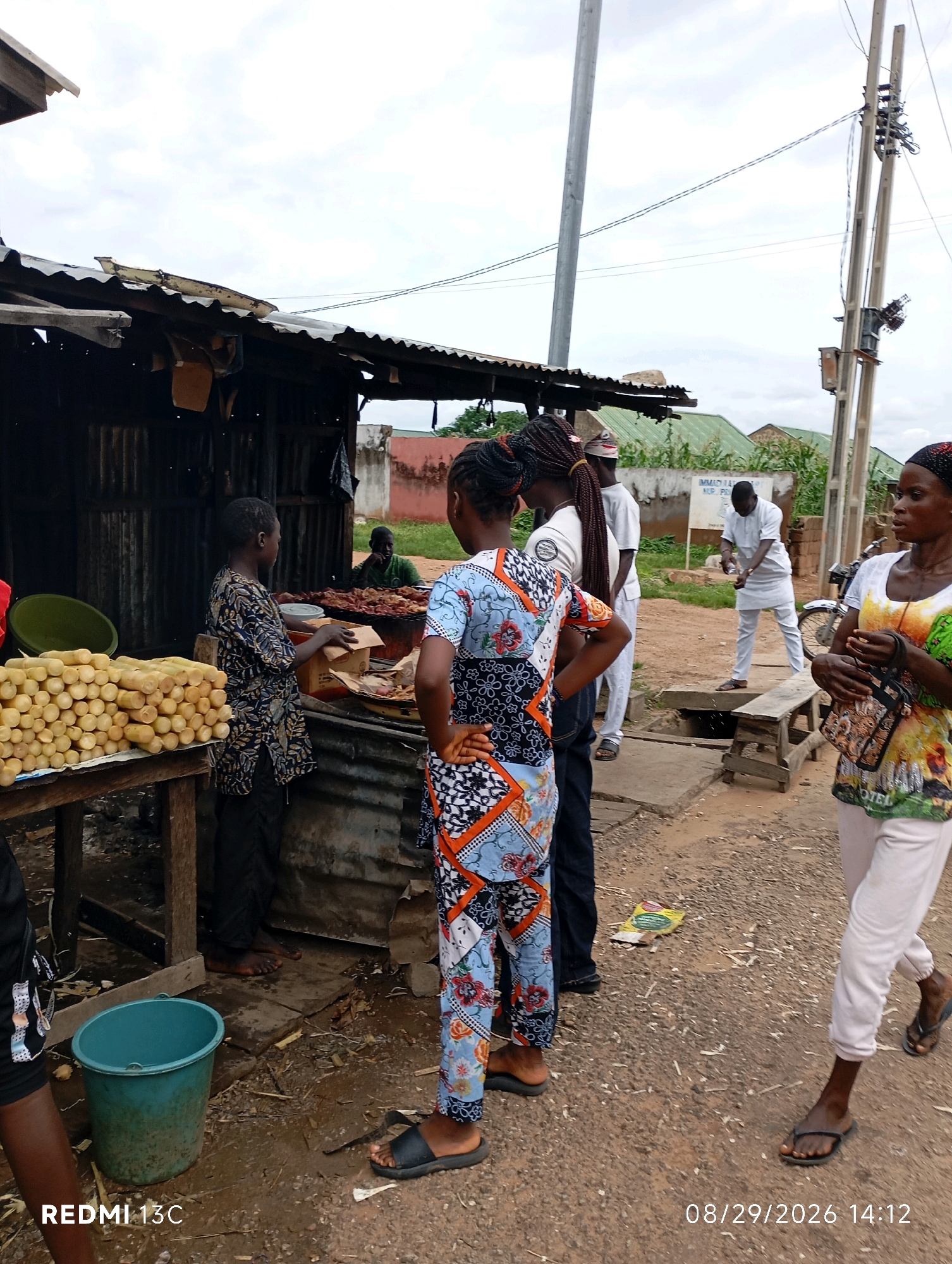The image depicts an outdoor market scene in Shendam, Nigeria. A food stall with a corrugated metal roof is visible on the left, displaying food items for sale, including what appears to be grilled meat and a large pile of corn cobs. Several individuals are present. A young person in a patterned shirt stands behind the stall, appearing to assist customers. Two women in colorful clothing are in the foreground, one facing away from the camera and the other walking to the right. In the background, a man in white attire is engaged in an activity, possibly related to a nearby drain or pit. Utility poles with electrical wires are prominent in the background, indicating the presence of infrastructure. The ground is dirt and appears wet, suggesting recent rain. The overall atmosphere is that of a typical busy market day.
