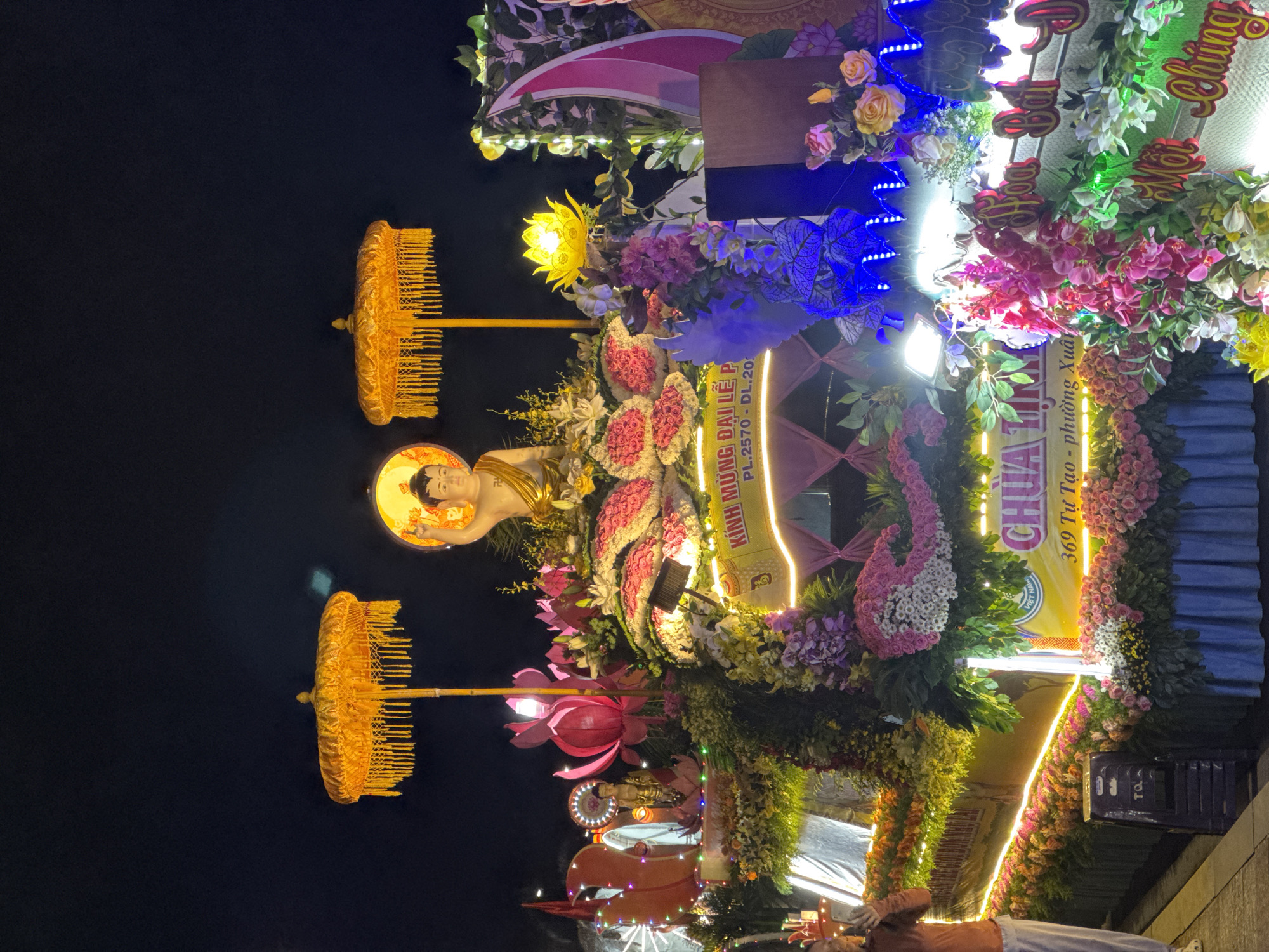 Captured in a vertical orientation, the image portrays a Buddhist parade float aglow at night. Dominating the frame is a golden statue of a Buddha figure, framed by a radiating halo. Accompanying the statue are two golden ornamental umbrellas, adding to the religious ambiance. The float is lavishly decorated with an abundance of flowers in varying shades of pink, yellow, and white, intertwined with lush green foliage. Blue and white LED lights are carefully woven into the display, enhancing the visual appeal. A yellow banner displays Vietnamese text, notably including "369 Tu Tao - phuong Xuan" which identifies the location as 369 Tu Tao Street in the Xuan neighborhood of Da Lat, Vietnam. A secondary, smaller Buddha statue is visible towards the bottom. The backdrop is a dark, clear night sky, emphasizing the luminance of the float. The float is positioned on a vehicle, indicated by the visibility of a person wearing orange clothing who is riding on it.